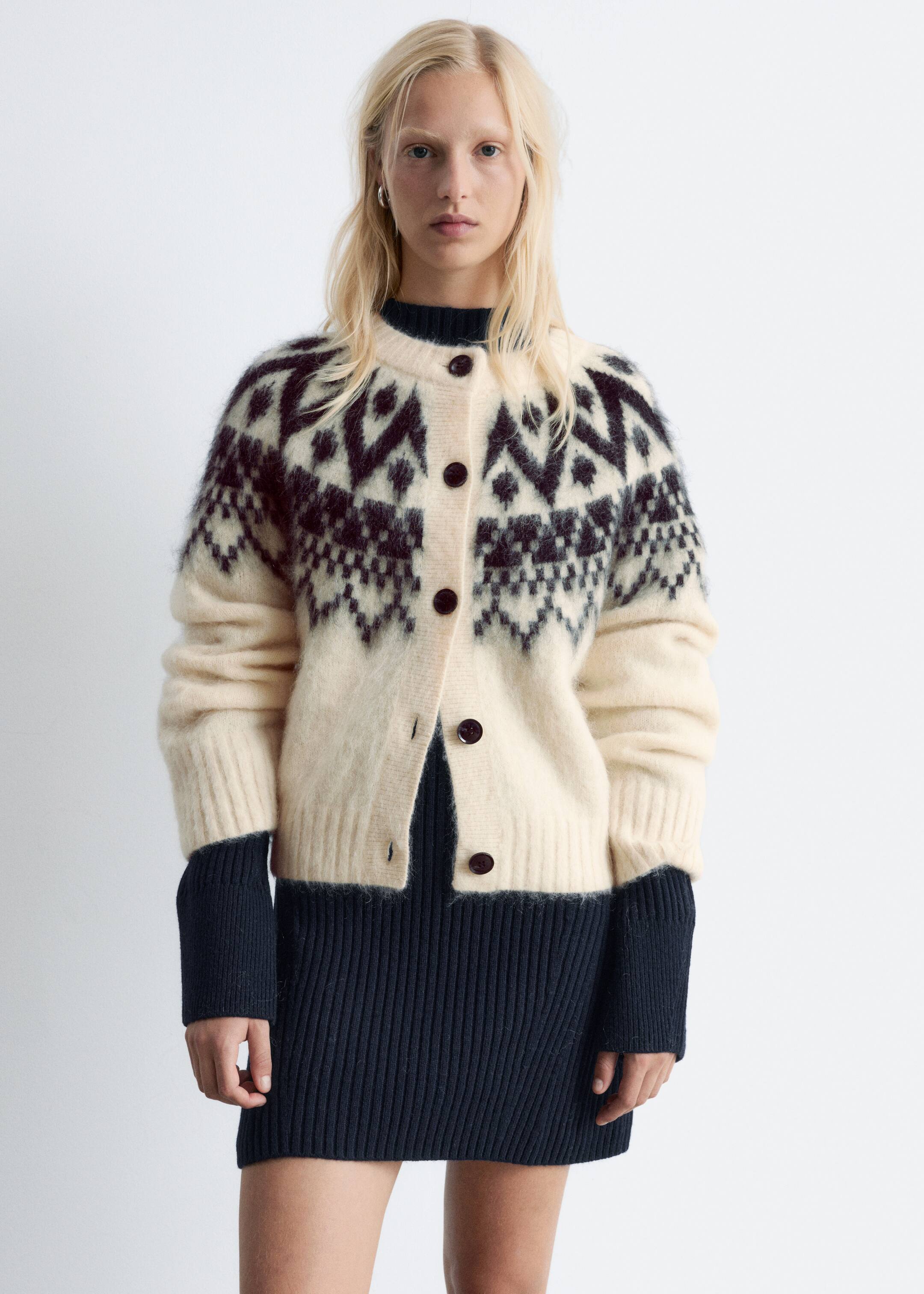 Größeres Bild anzeigen: Eine cremefarbene Strickjacke mit einem dunklen Jacquard-Muster über Brust und Schultern wird vom Model getragen. Die kastenförmige Strickjacke hat gerippte Bündchen und einen gerippten Saum, fünf dunkle Knöpfe vorne, die teilweise geschlossen sind, und wird über einem dunklen, gerippten Kleidungsstück getragen.