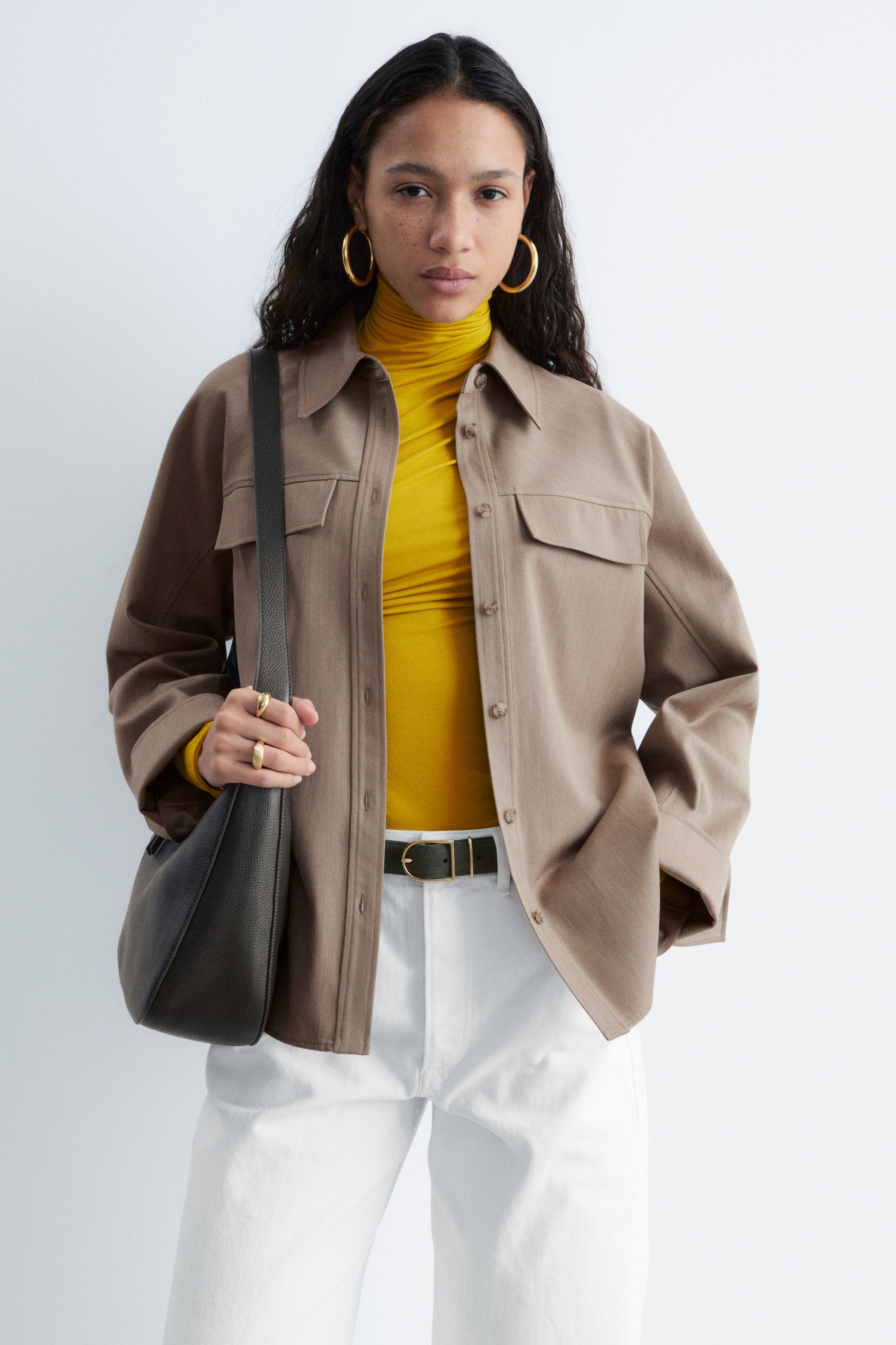 Visualizza immagine più grande: La giacca camicia marrone presenta un colletto a punta, una chiusura frontale con bottoni e due tasche sul petto con patta e chiusura a bottone. È indossata aperta sopra un dolcevita giallo, mettendo in risalto la sua vestibilità rilassata e le maniche lunghe con polsini abbottonati.
