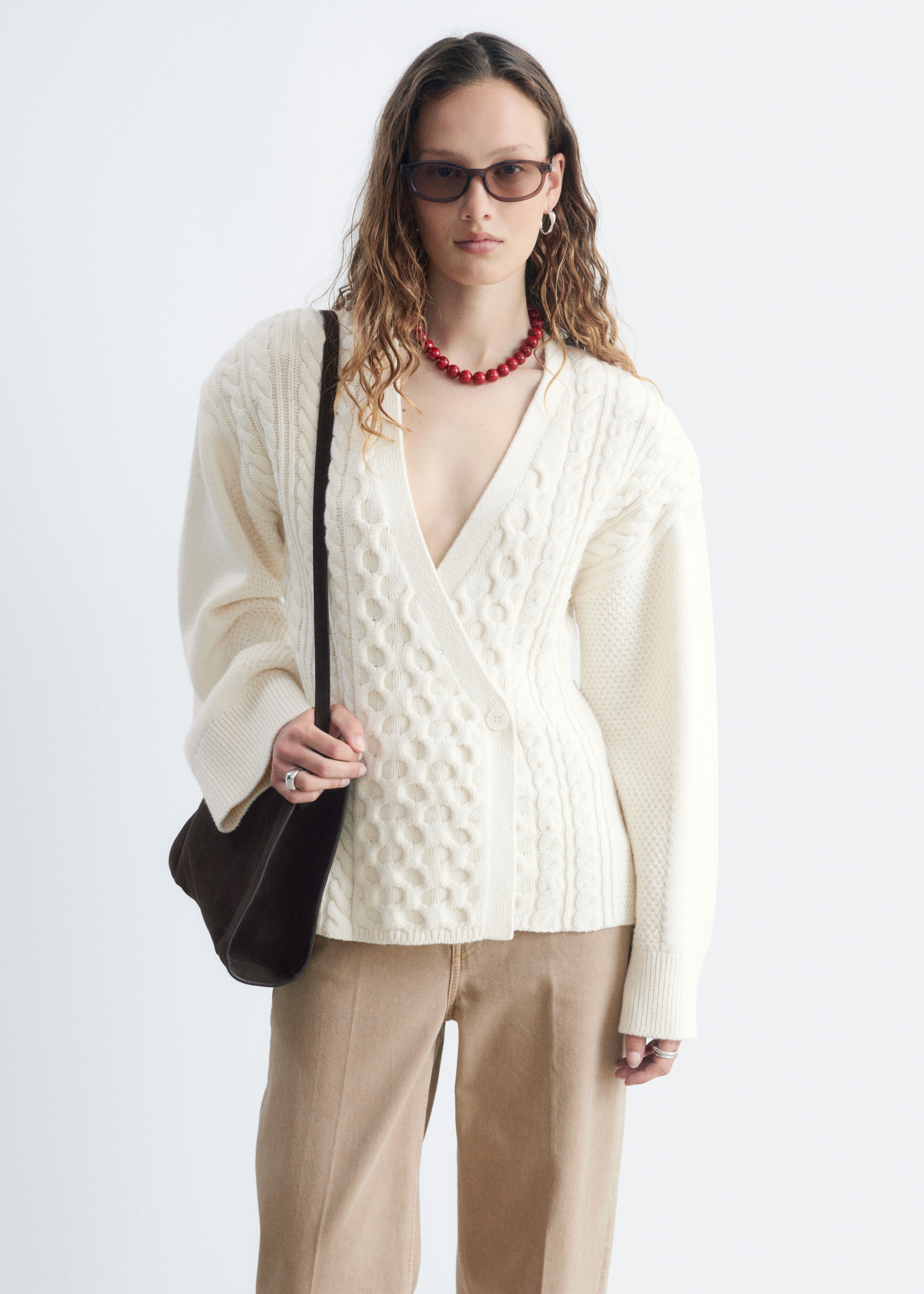 Um cardigan de mistura de lã e algodão de cor creme está deitado plano, com a frente voltada para cima. Apresenta um design envolvente com fecho de botão lateral, exibindo uma mistura de padrões de malha de trança e bolinhas na parte da frente e malhas de textura fina nos painéis laterais. As mangas terminam com punhos largos canelados.