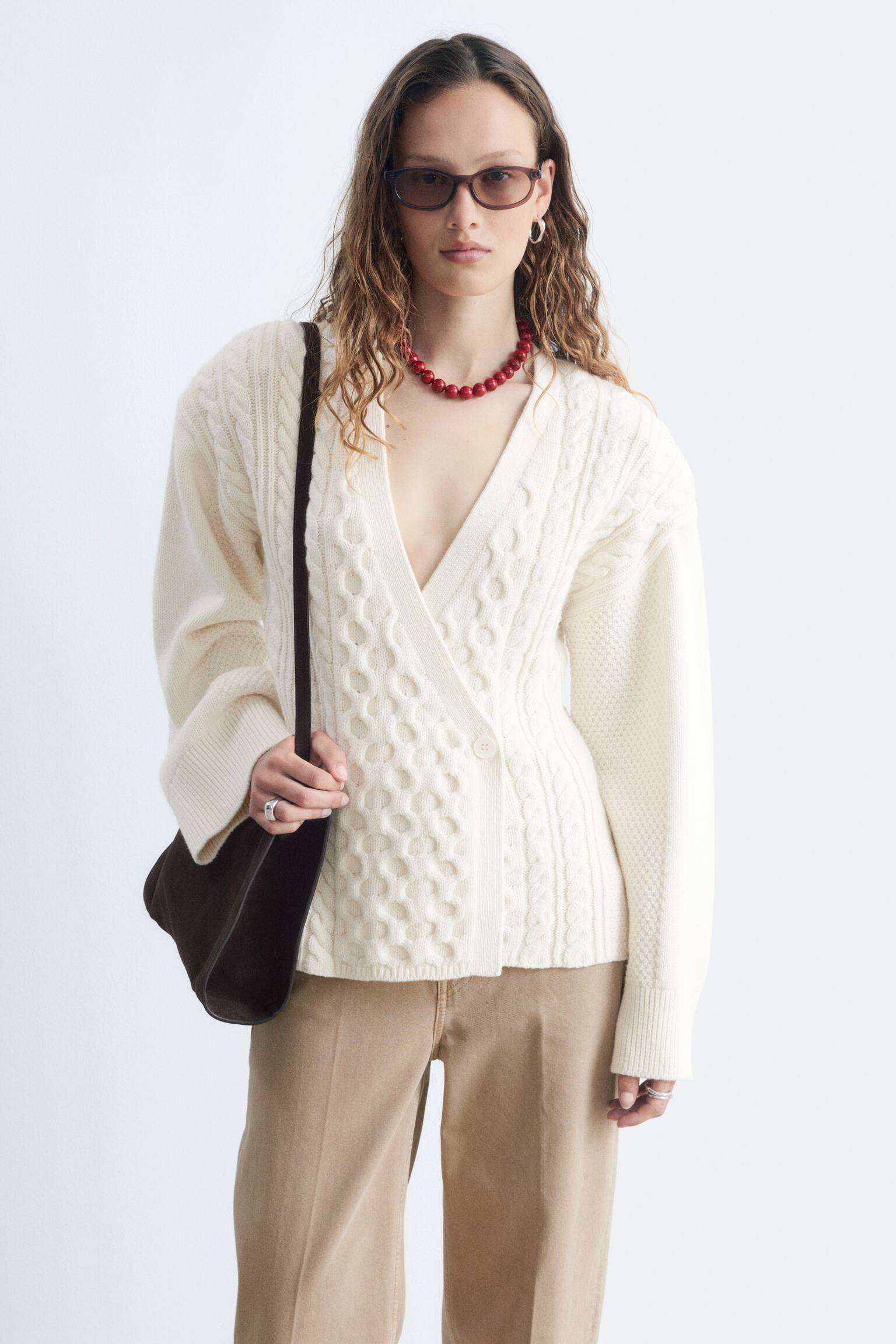 En cremefarvet kabelstrikket slå-om cardigan bæres, der viser en dyb V-udskæring og en enkelt knaplukning i taljen. Den har forskellige kabelstrikmønstre, herunder bobler og snoede designs, med ribbede manchetter og en ribbet kant, mens en mørk taskestrop krydser over dens venstre skulder. - 1