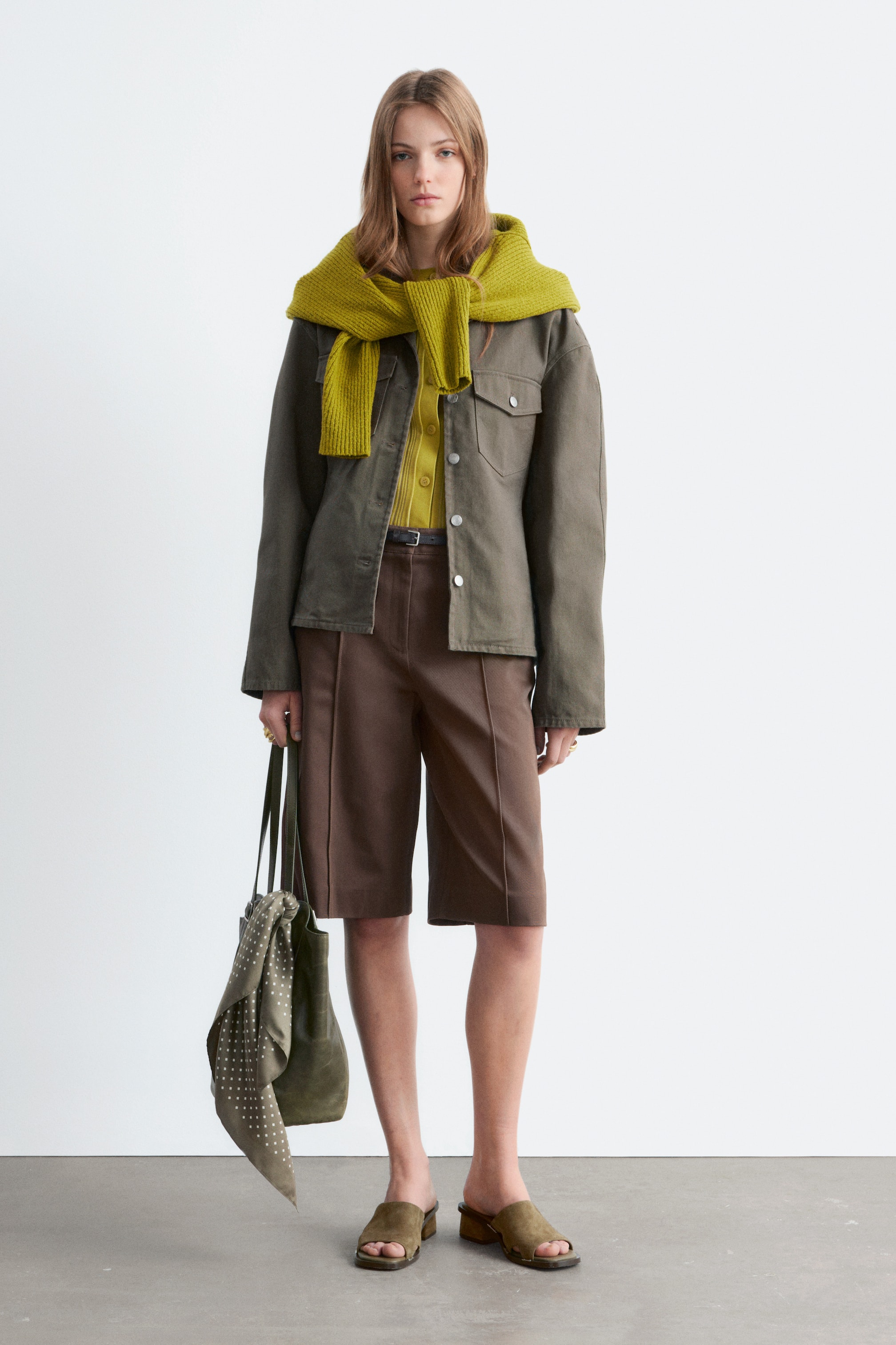 Visualizza immagine più grande: Una giacca utility verde oliva scuro con tasche sul petto viene indossata aperta sopra un cardigan giallo a coste, con un maglione a maglia grossa verde brillante drappeggiato sulle spalle. Pantaloncini marroni al ginocchio con cuciture frontali sono abbinati a una cintura sottile scura, una borsa a tracolla verde oliva con una sciarpa a pois e sandali con tacco a blocco aperti verde oliva, tutti presentati su un modello rivolto in avanti.
