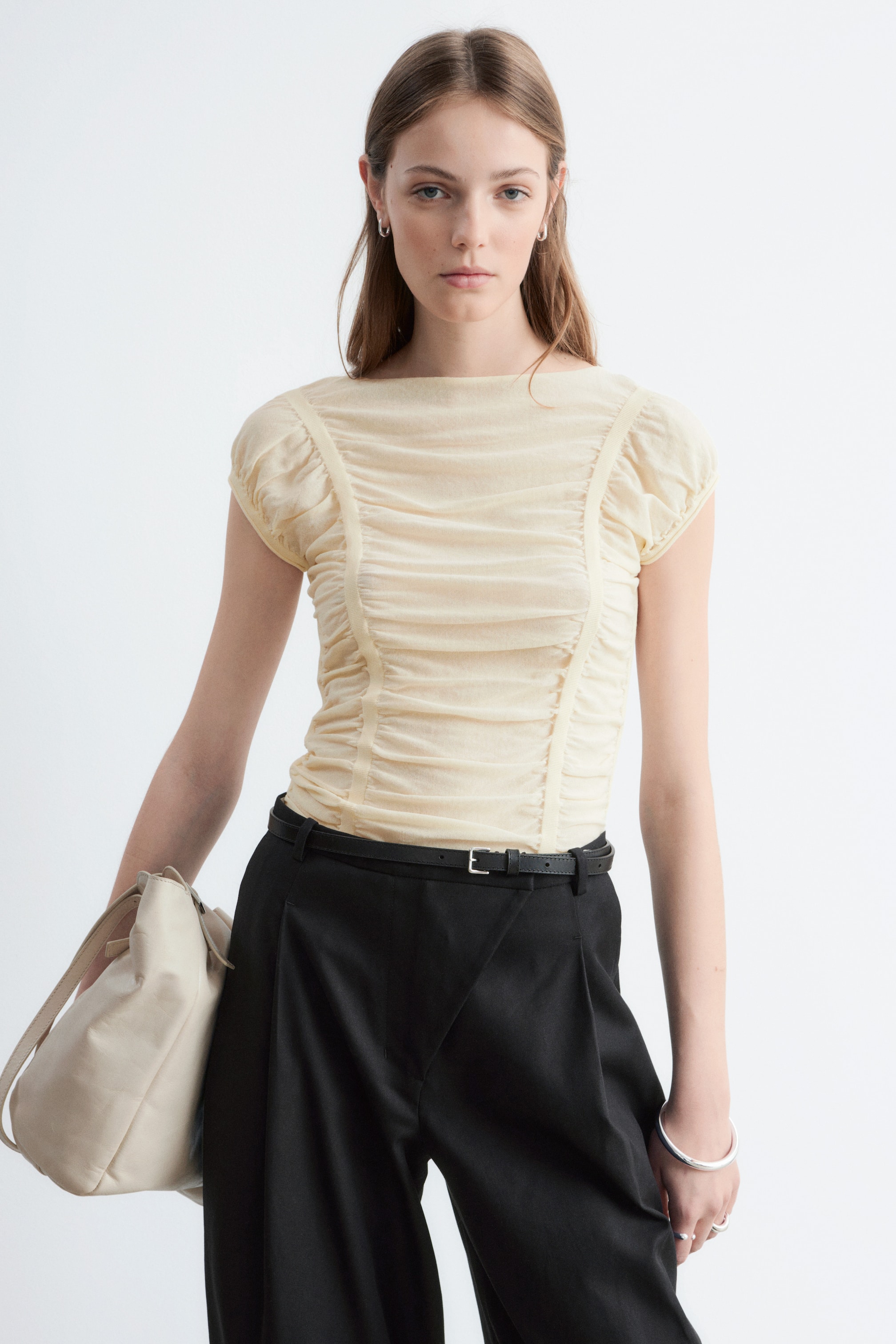 Grotere afbeelding bekijken: Een lichtgele gerimpelde top wordt gepresenteerd met de voorkant naar voren gericht. Het heeft een boothals en korte geplooide kapmouwtjes, en de aansluitende body toont uitgebreide verticale en horizontale rimpelingen, gescheiden door gladde verticale panelen.