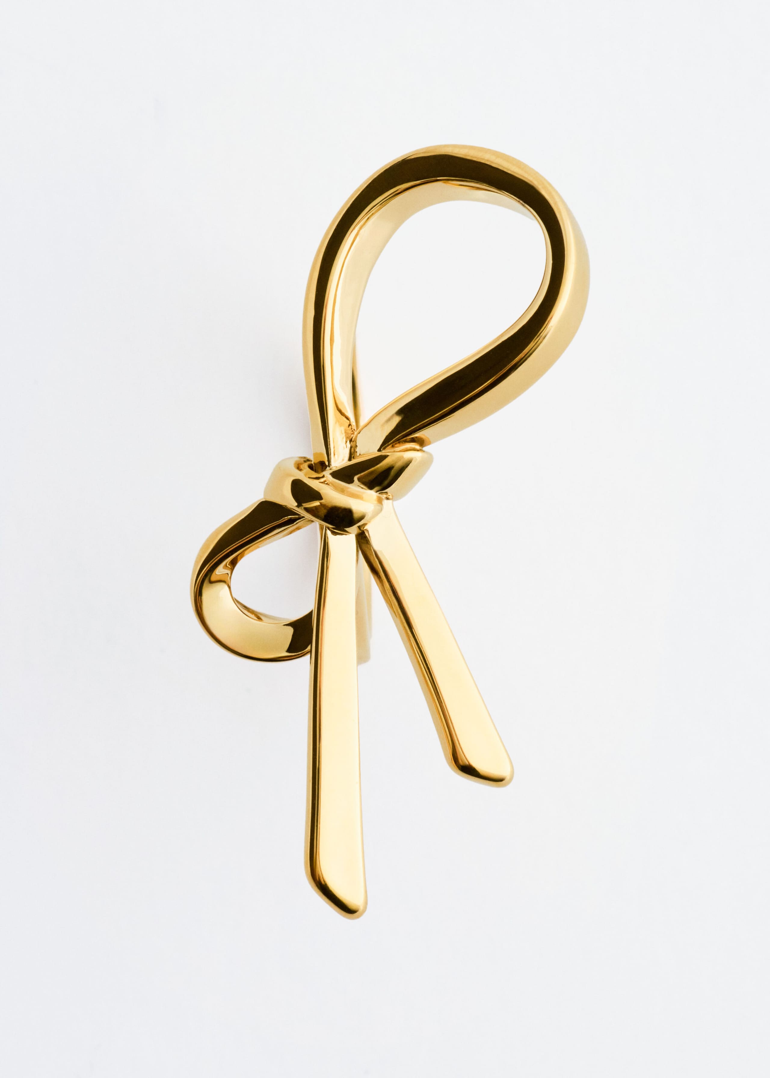 A polished brass ring displays a shiny, reflective gold finish, featuring a tied ribbon knot design with two hanging ends seen from the front.