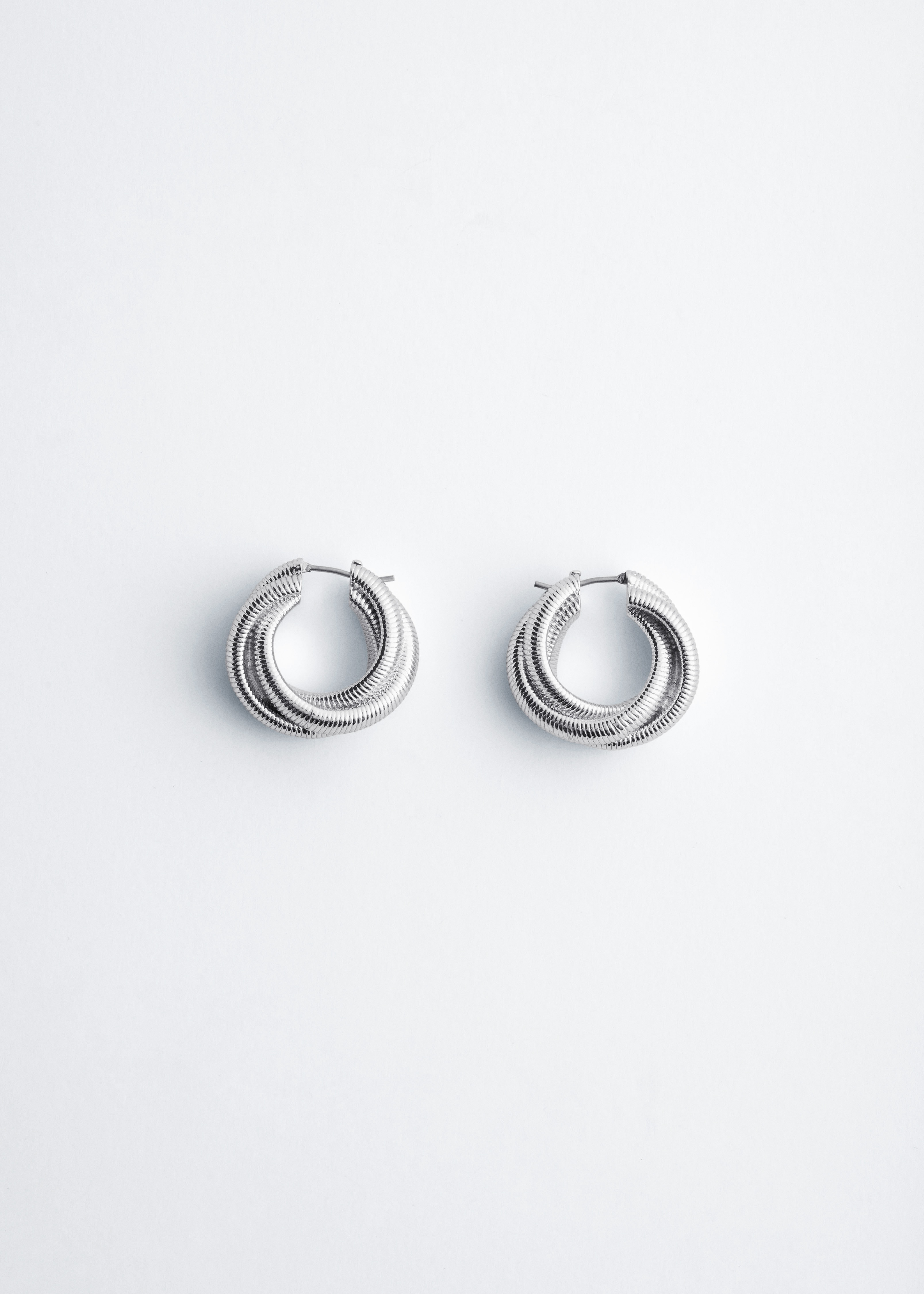A black wool-cotton knitted vest featuring a ribbed V-neck is worn by the model over a white collared shirt. A silver-toned plated brass hoop earring with a twisted, textured surface is also worn, visible on the modelâs ear.