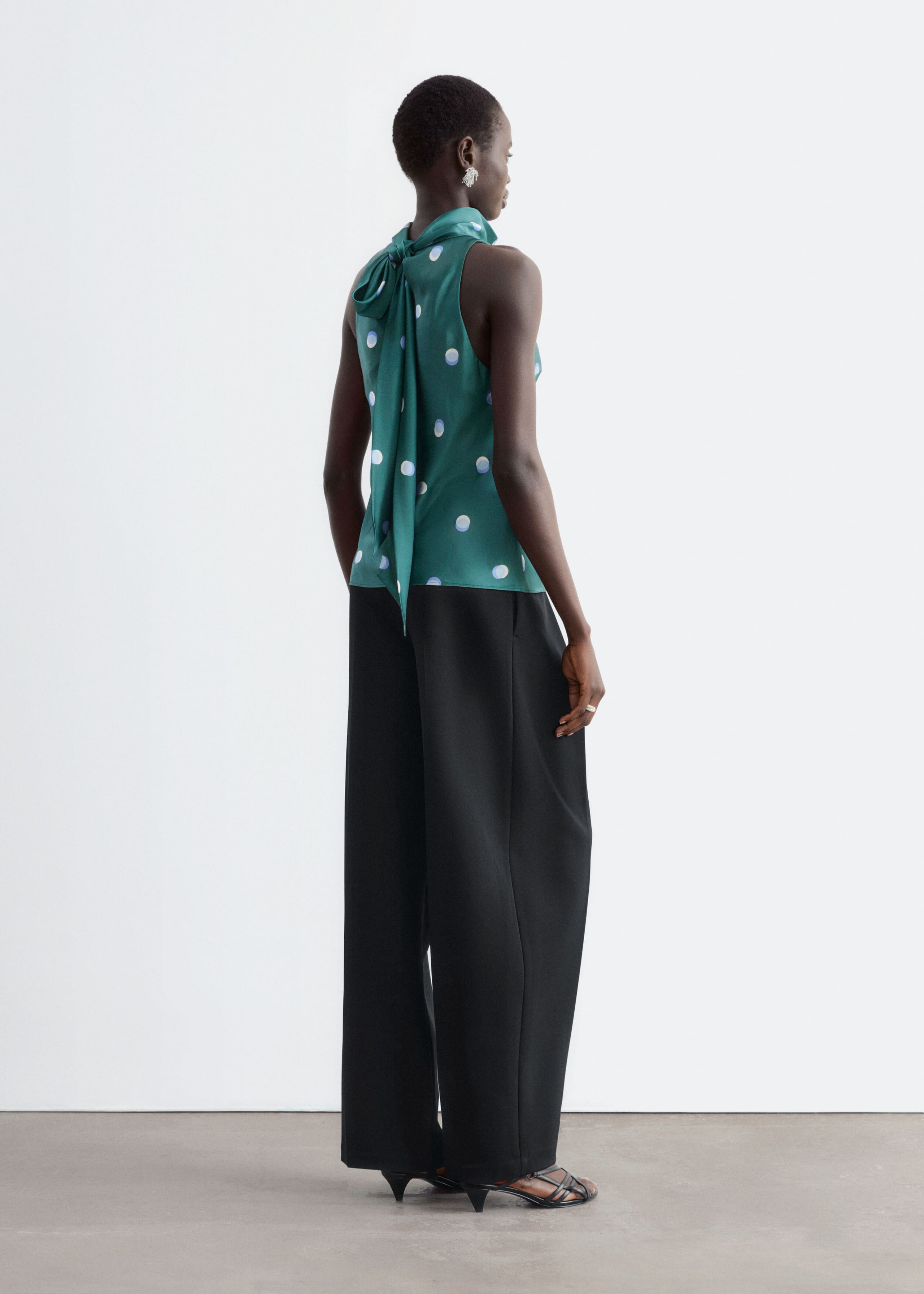 A green satin sleeveless top with a light blue and white polka dot pattern is being worn by the model, showcasing the back view. The top features a high neckline with a long scarf tied at the back that drapes down, and is styled with black wide-leg trousers.
