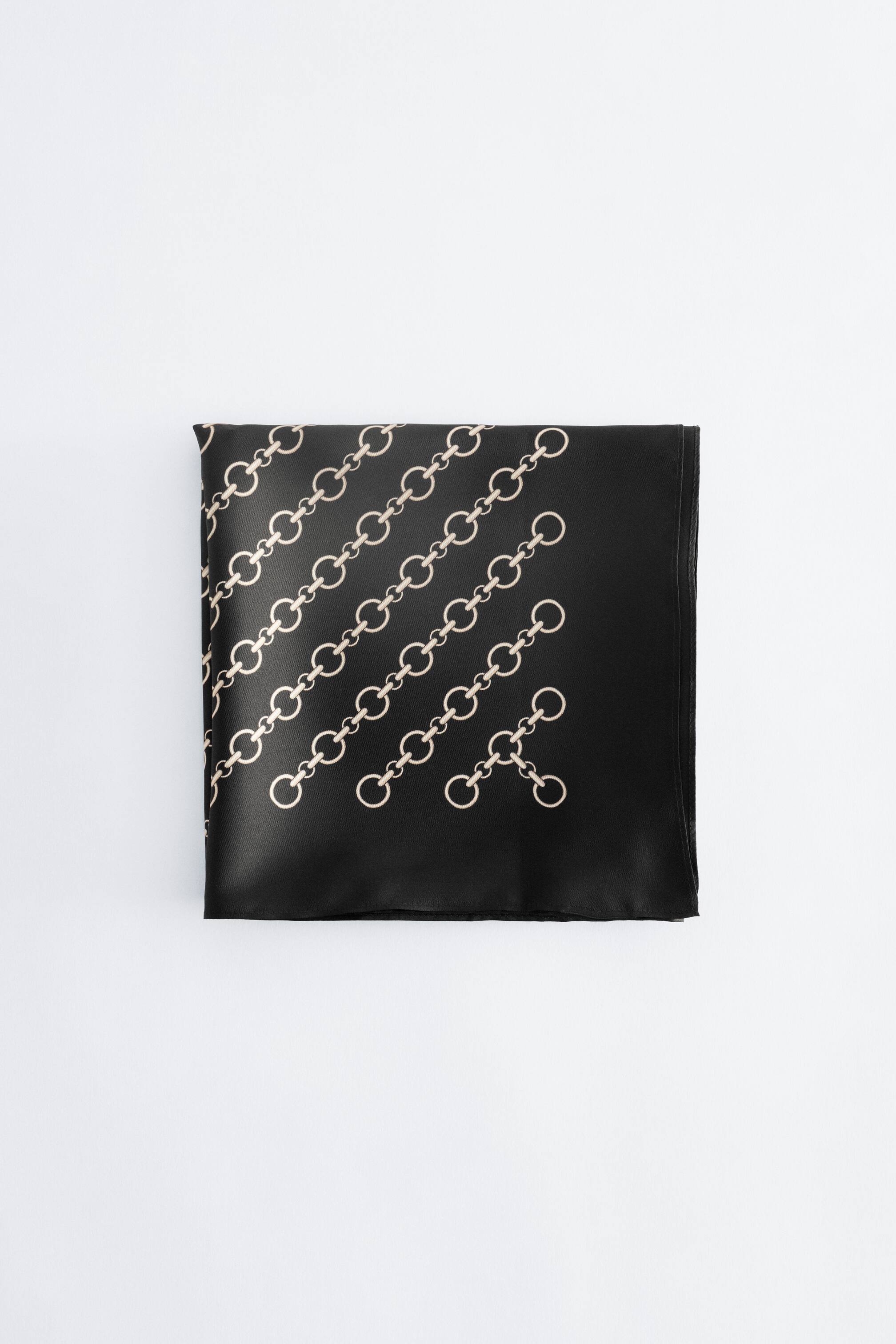 Visualizza immagine più grande: Una sciarpa di seta nera è mostrata piegata a quadrato, con un motivo diagonale di maglie a catena beige sulla sua superficie liscia con finitura satinata.