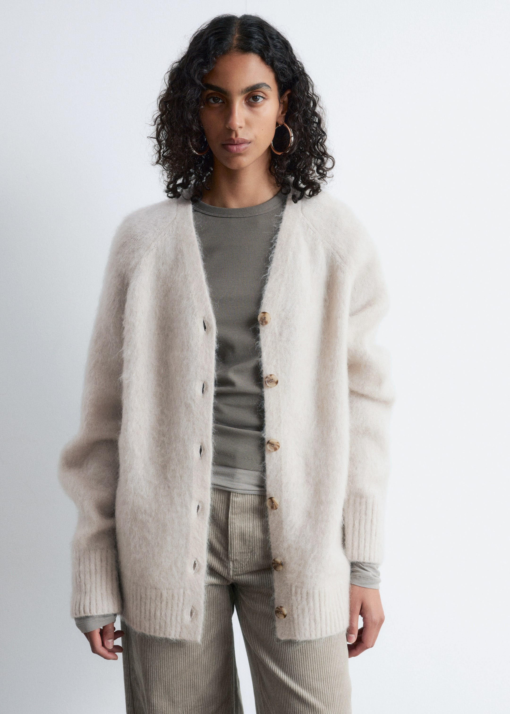 Visualizza immagine più grande: Il Cardigan Lungo in Misto Mohair è indossato dalla modella, mostrando una texture pelosa color beige chiaro, scollo a V, maniche raglan, polsini e orlo a coste. È abbinato aperto, con evidenti bottoni marroni lungo il davanti, sovrapposto a un top a maniche lunghe a coste.