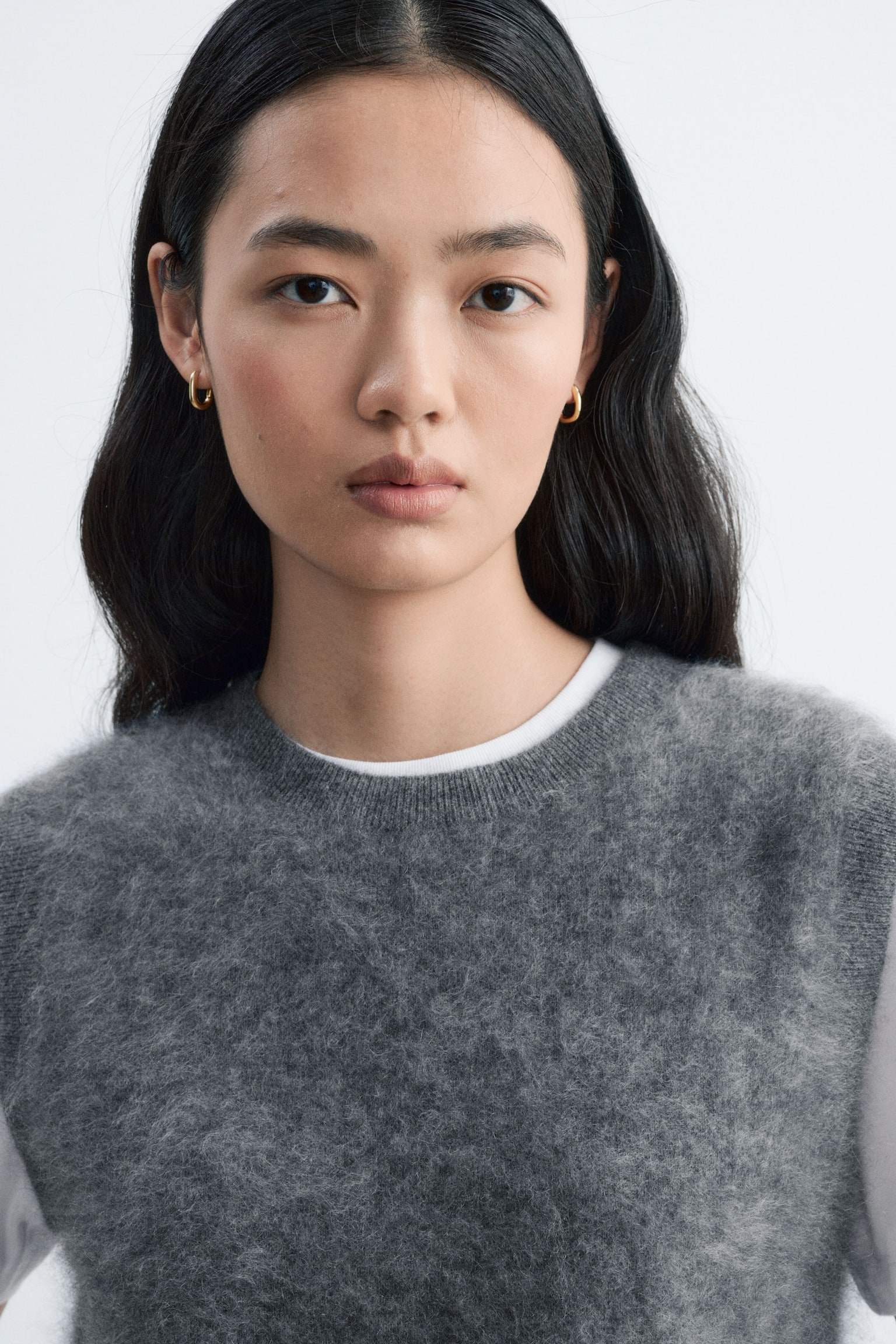 Un chaleco de cashmere jaspeado gris, tejido de punto, con cuello redondo acanalado, lo lleva la modelo sobre una camiseta blanca, destacando su textura suave y los remates acanalados de las sisas. - 1