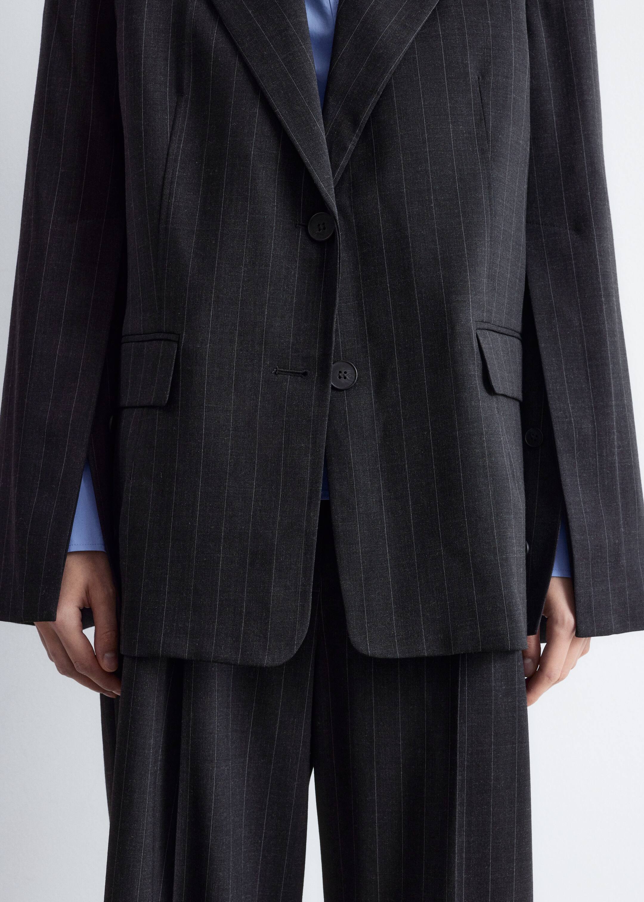 Größeres Bild ansehen: Ein anthrazitfarbenes Nadelstreifenanzug wird getragen, mit einem einreihigen Blazer mit Kerbrevers, einem Zweiknopfverschluss mit geschlossenem unteren Knopf und zwei vorderen Pattentaschen. Der locker sitzende Blazer ist über einem blauen Hemd geschichtet, und passende weite Hosen sind ebenfalls sichtbar.