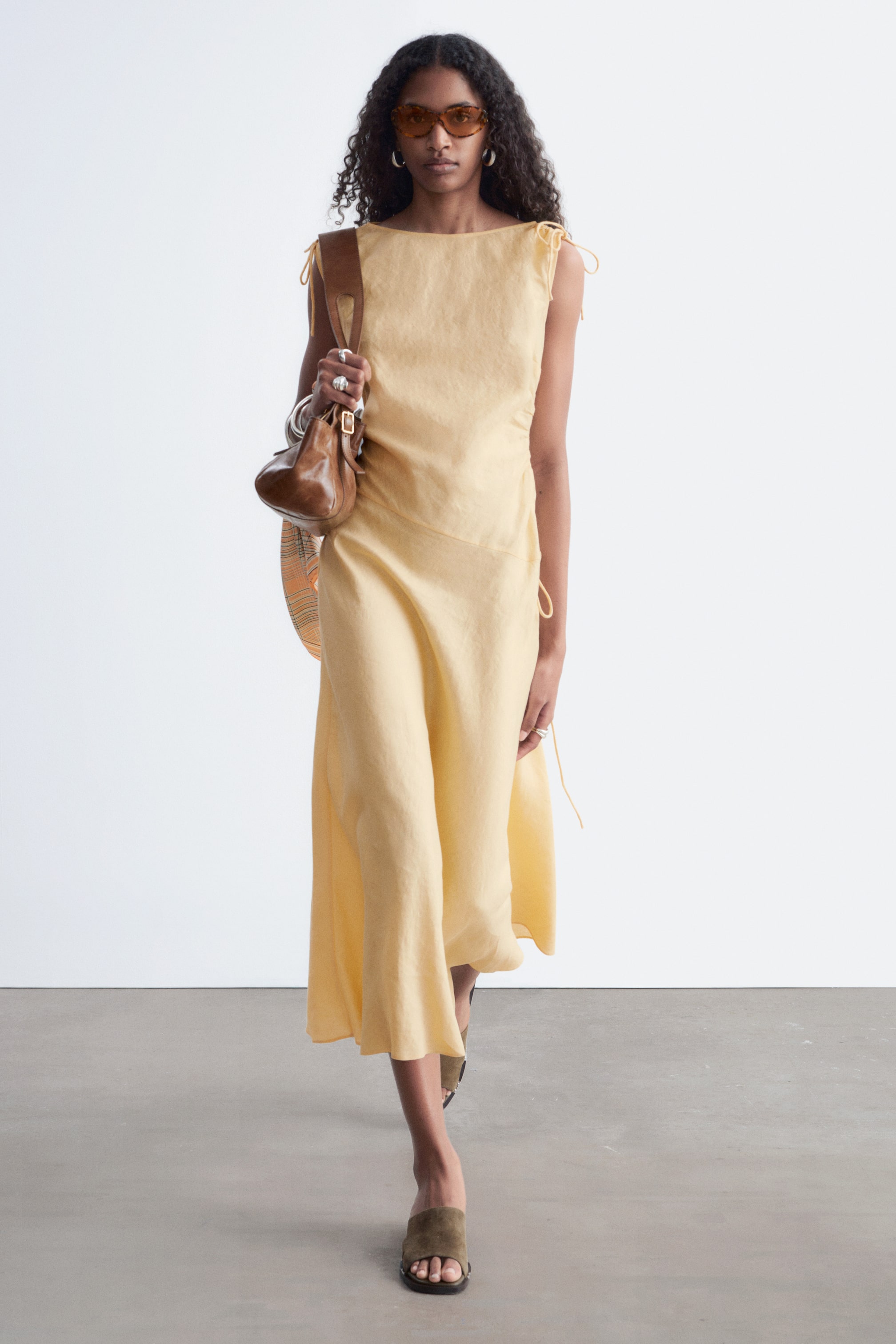 Agrandir l'image: Une robe sans manches jaune pâle présente un large décolleté et des détails noués sur les épaules, tandis qu'un corsage ajusté se transforme en une jupe mi-longue fluide avec un panneau de style portefeuille et un accent noué sur le côté, se drapant élégamment lorsque la personne avance.