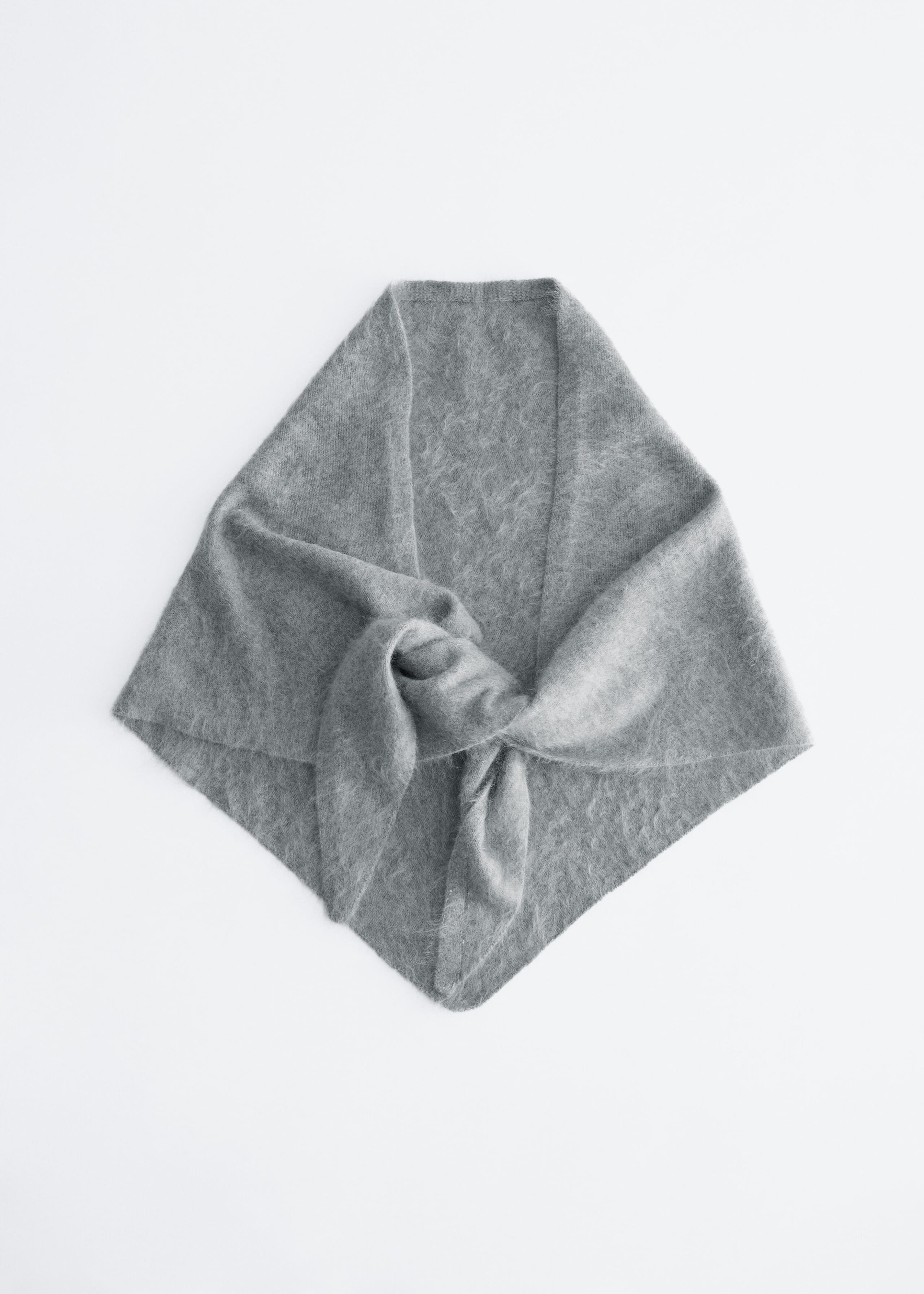 A gray knit scarf with a fuzzy, brushed texture is lying flat, showcasing its triangular shape from the front. The design is styled with its ends loosely knotted together in the center, highlighting its soft drape and mélange finish.