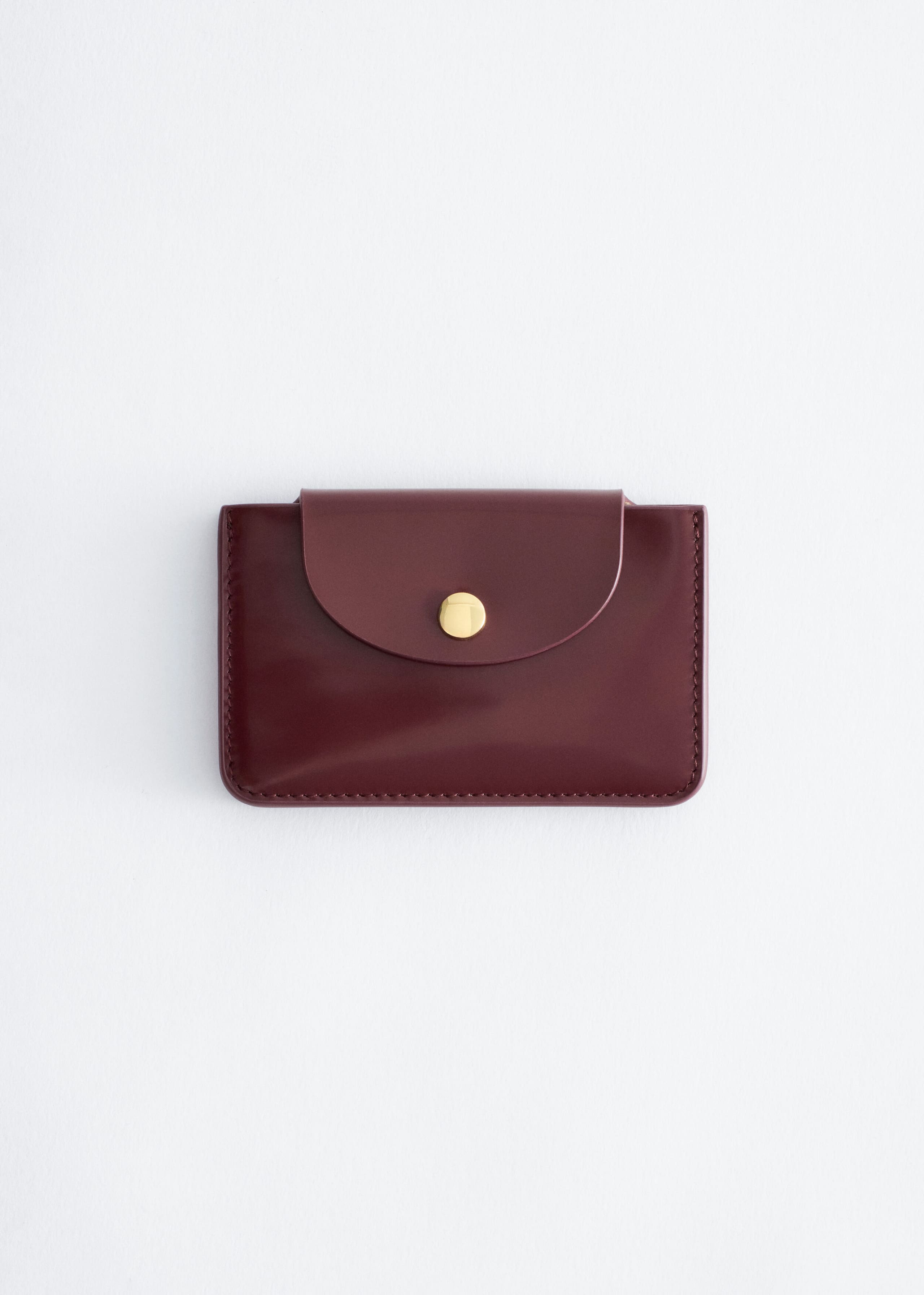 A burgundy leather cardholder is shown from the front, featuring a smooth finish, tonal stitching along the edges, and a curved fold-over flap with a gold-tone snap closure.