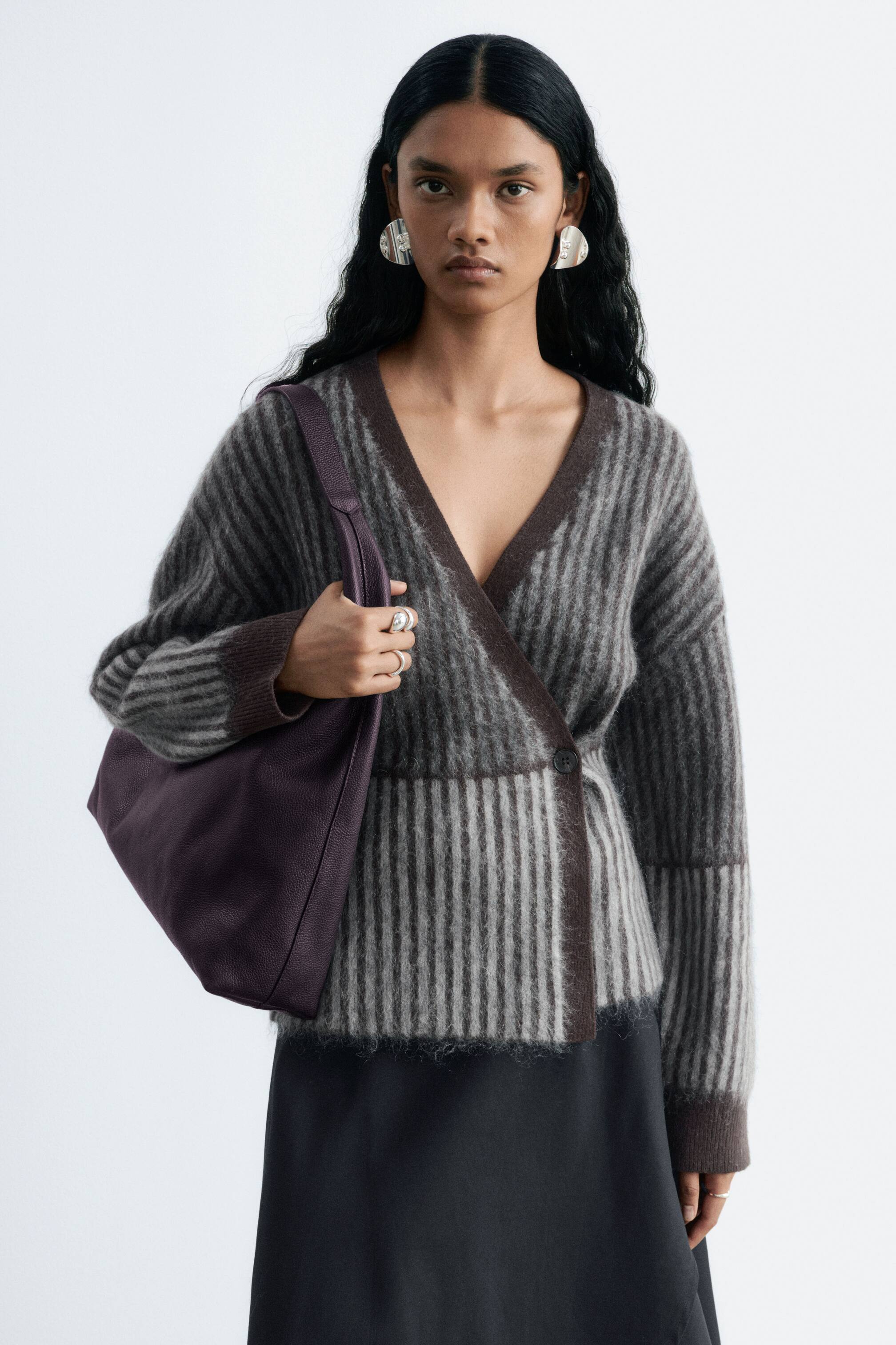 Vezi imaginea mai mare: O geantă hobo din piele mov cu textură granulată este ținută în mâna dreaptă a modelului și purtată pe umăr. Modelul poartă, de asemenea, un cardigan petrecut cu decolteu în V, cu dungi verticale maro și gri deschis, având o textură tricotată pufoasă, purtat peste o fustă neagră.