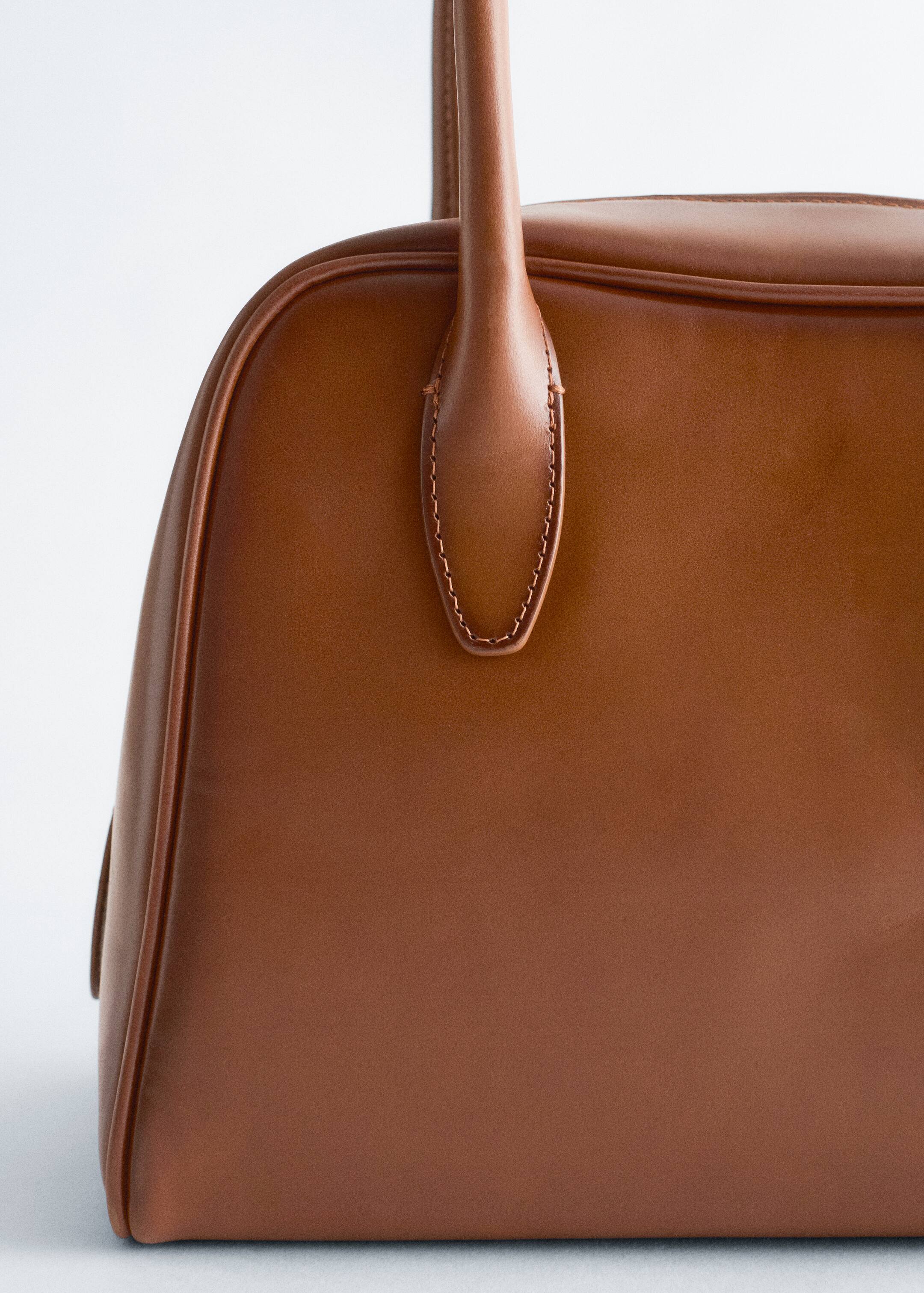 Größeres Bild ansehen: Eine braune Handtasche zeichnet sich durch eine glatte, strukturierte Oberfläche mit Paspeldetails entlang ihrer abgerundeten Kanten aus. Ein schlanker oberer Griff wird durch einen verlängerten, genähten Riemen befestigt.