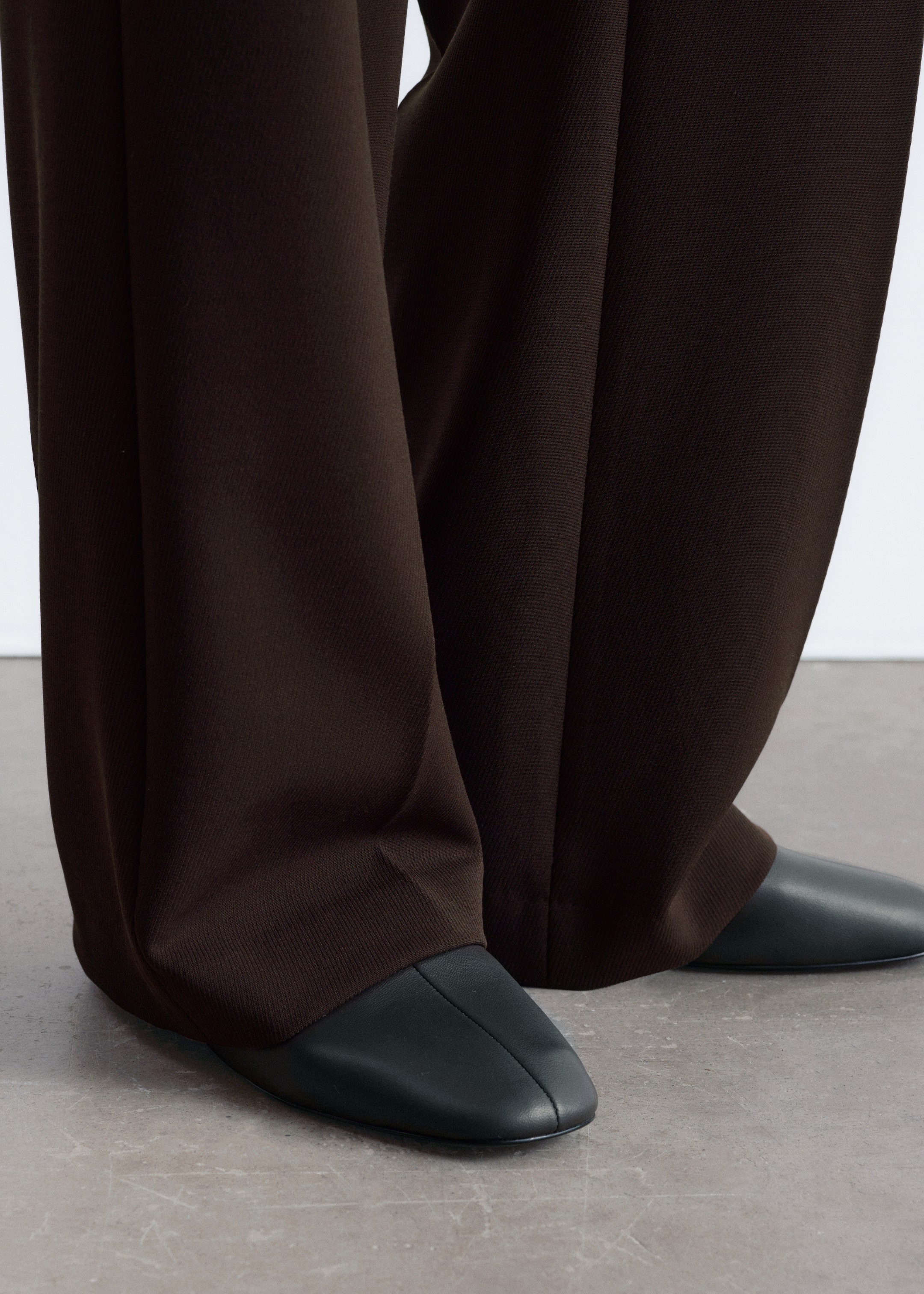 Agrandir l'image: Un pantalon marron foncé à jambes larges tombe sur des chaussures plates vert foncé à bout carré. Le pantalon présente une texture subtile de tissage en diagonale, tandis que les chaussures, légèrement inclinées vers l'extérieur, arborent une couture centrale sur leurs boîtes à orteils.