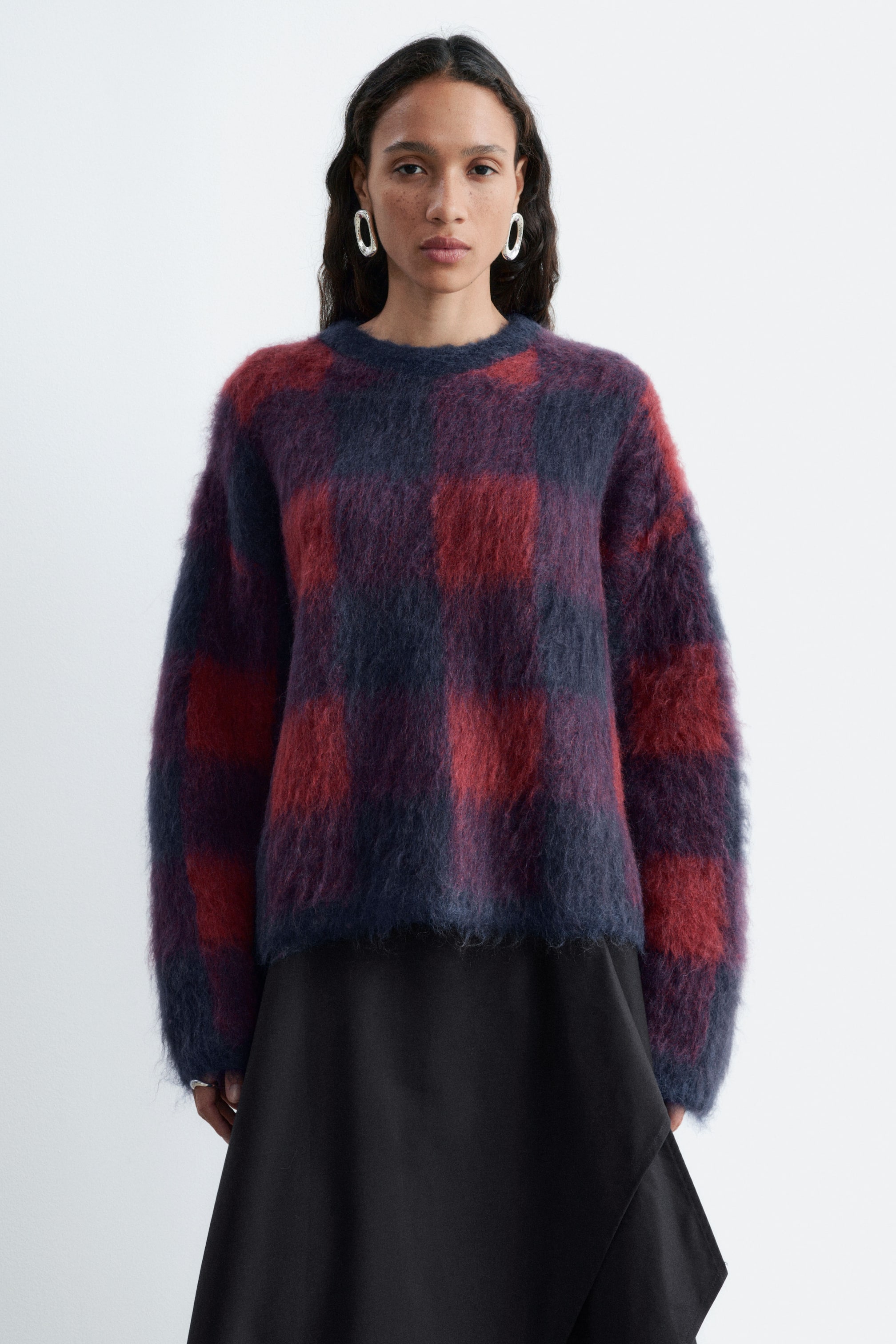 Größeres Bild anzeigen: Ein flauschiger Pullover aus Mohair-Mischung mit rotem und dunkelblauem Karomuster wird vom Model getragen. Der Rundhalspullover verfügt über lange Ärmel und eine lockere Passform und ist über einem schwarzen Rock gestylt.