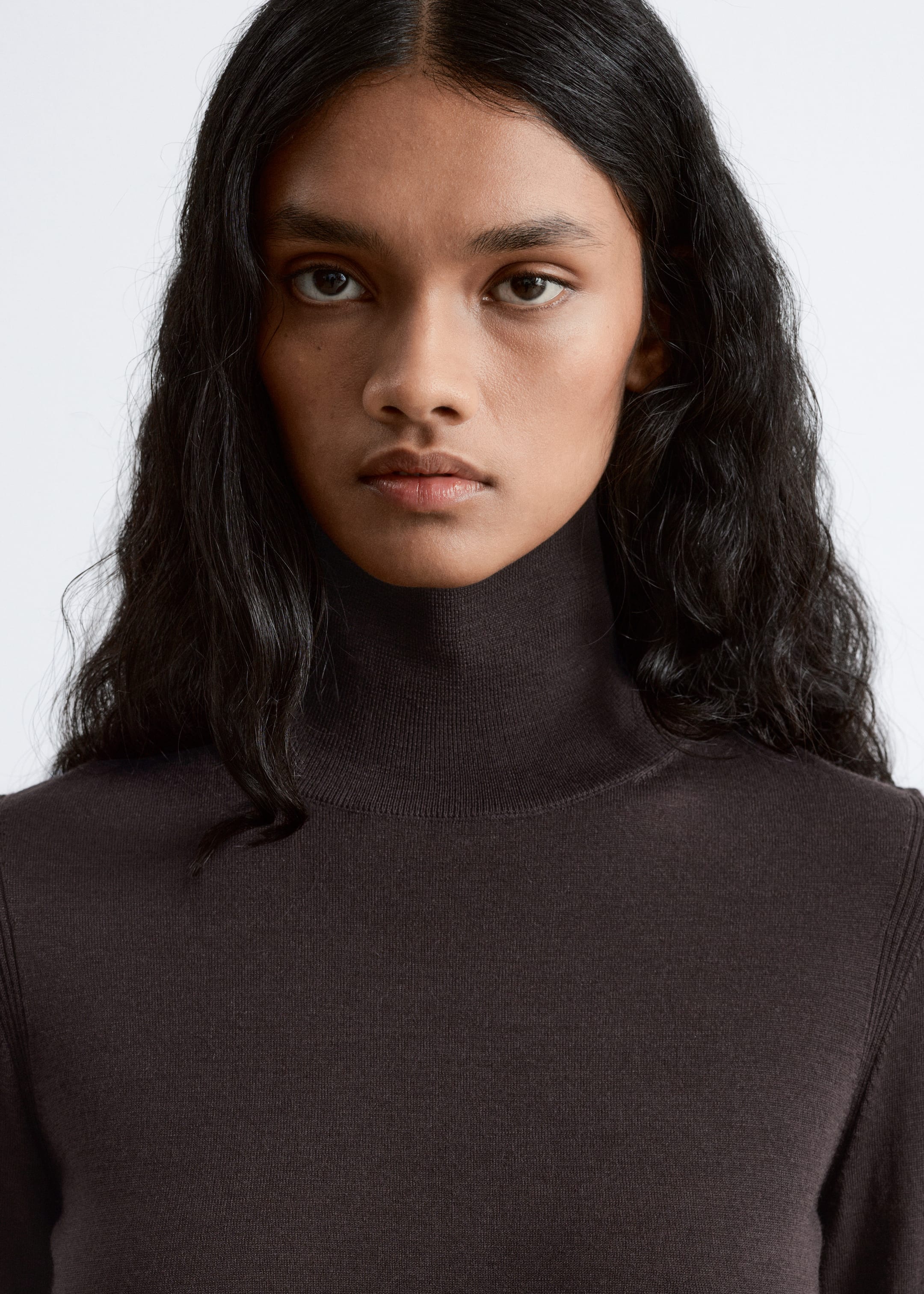 Grösseres Bild ansehen: Ein dunkelbrauner Rollkragenpullover wird von vorne gezeigt, mit einem eng anliegenden, hohen Kragen. Sein glattes Strickgewebe enthält subtile vertikale Rippendetails, die entlang der Seite des Kleidungsstücks sichtbar sind.