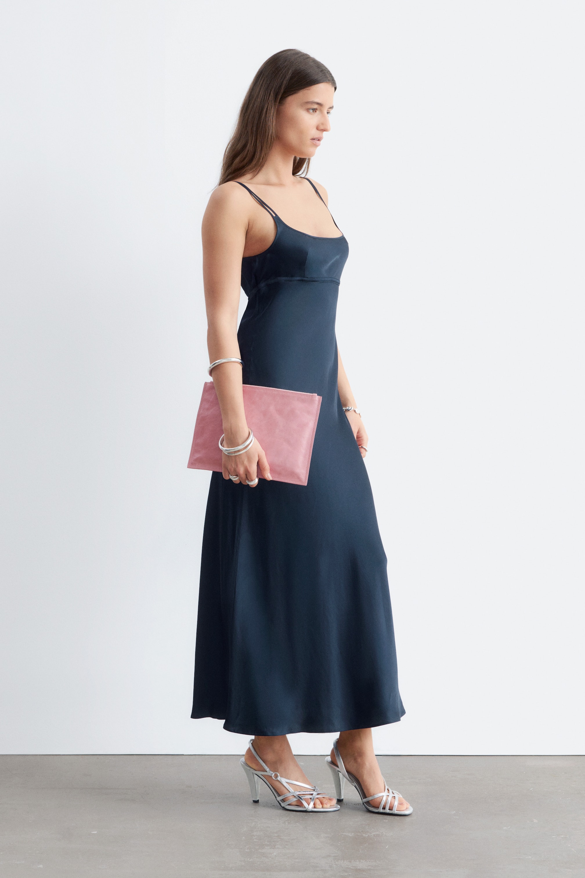 Agrandir l'image: Une robe midi en satin bleu foncé, présentant une silhouette élancée, un décolleté carré avec des doubles bretelles et un biais plat au niveau de la poitrine, est portée par le mannequin. Le mannequin tient une pochette rose et porte des talons argentés à brides ainsi que des bracelets.
