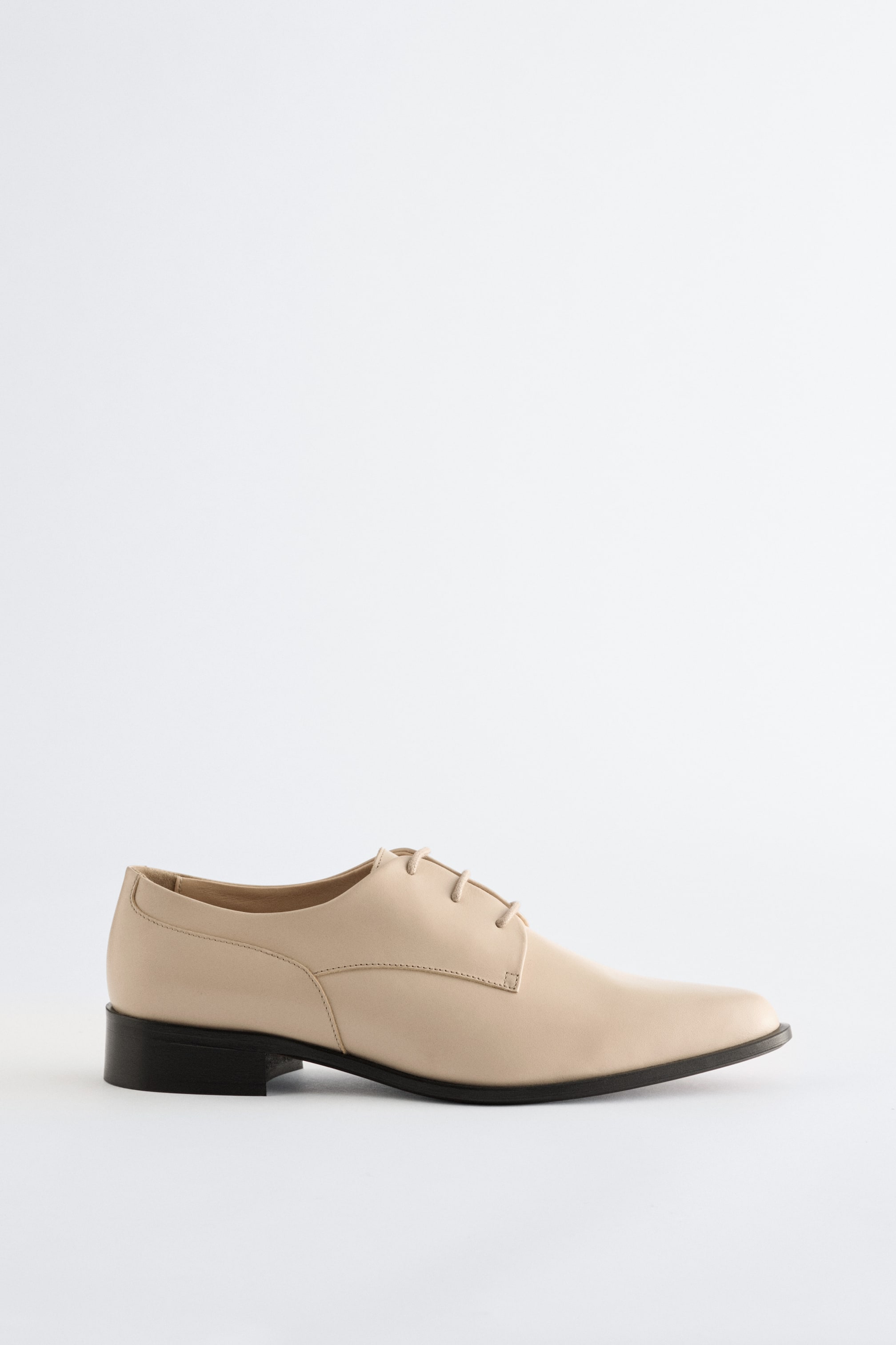 Agrandir l'image: Une chaussure Derby en cuir beige clair est présentée en profil latéral, avec un bout pointu et un talon bloc bas noir. Elle affiche une tige lisse et un système de laçage ouvert avec quatre paires de lacets assortis.