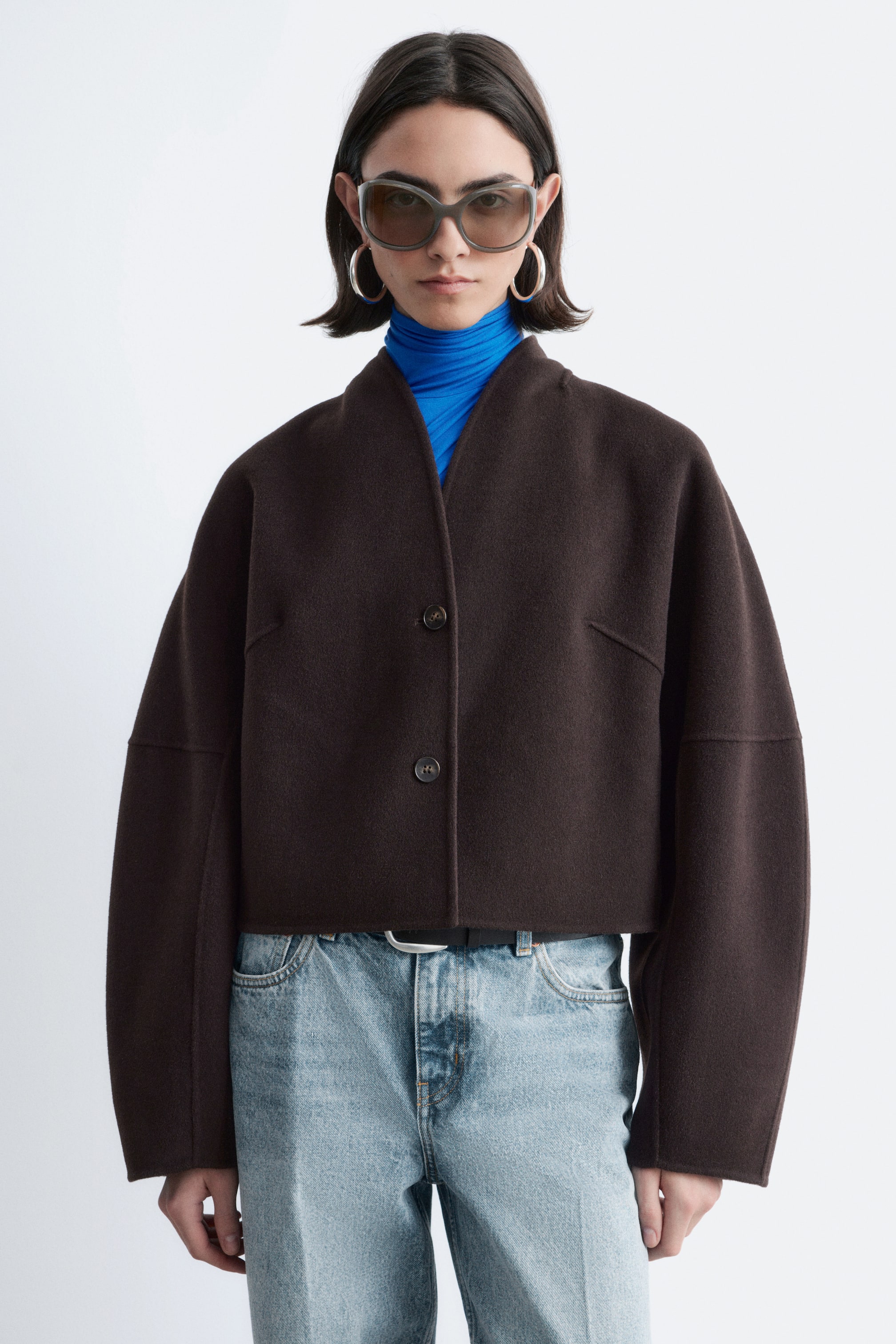 Se større billede: En mørkebrun cropped jakke fremviser en kraveløs V-udskæring og to frontknapper. Den har brede, lange ærmer med vandrette sømdetaljer, lagdelt over en blå turtleneck.