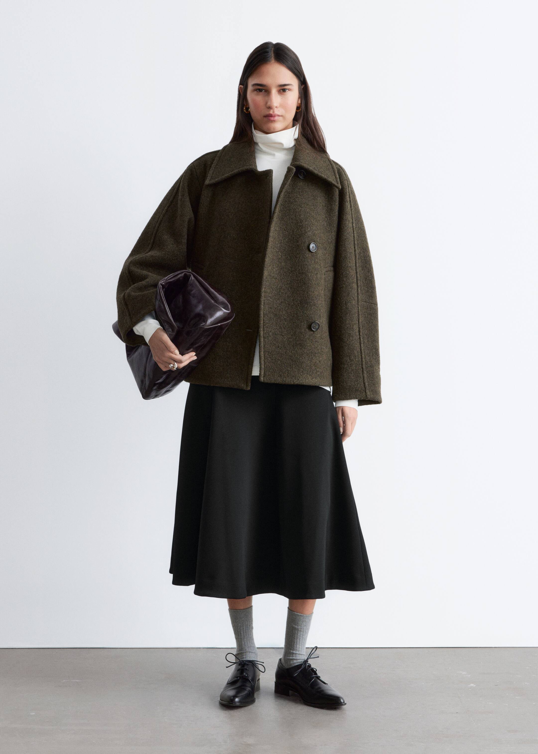 Se større billede: En mørk olivengrøn, tekstureret, boxy, kort jakke præsenteres forfra, med brede notch revers og en dobbeltradet front med to synlige mørke knapper. Den er stylet åben, hvilket afslører en hvid turtleneck nedenunder, og har brede ærmer.