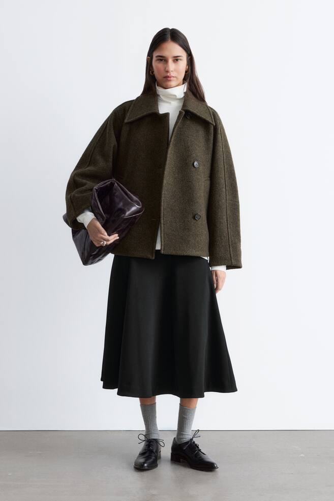 Une veste courte et texturée de couleur vert olive foncé, à la coupe carrée, est présentée de face, avec de larges revers crantés et un devant croisé à double boutonnage avec deux boutons sombres visibles. Elle est portée ouverte, révélant un col roulé blanc en dessous, et possède des manches larges. - 1