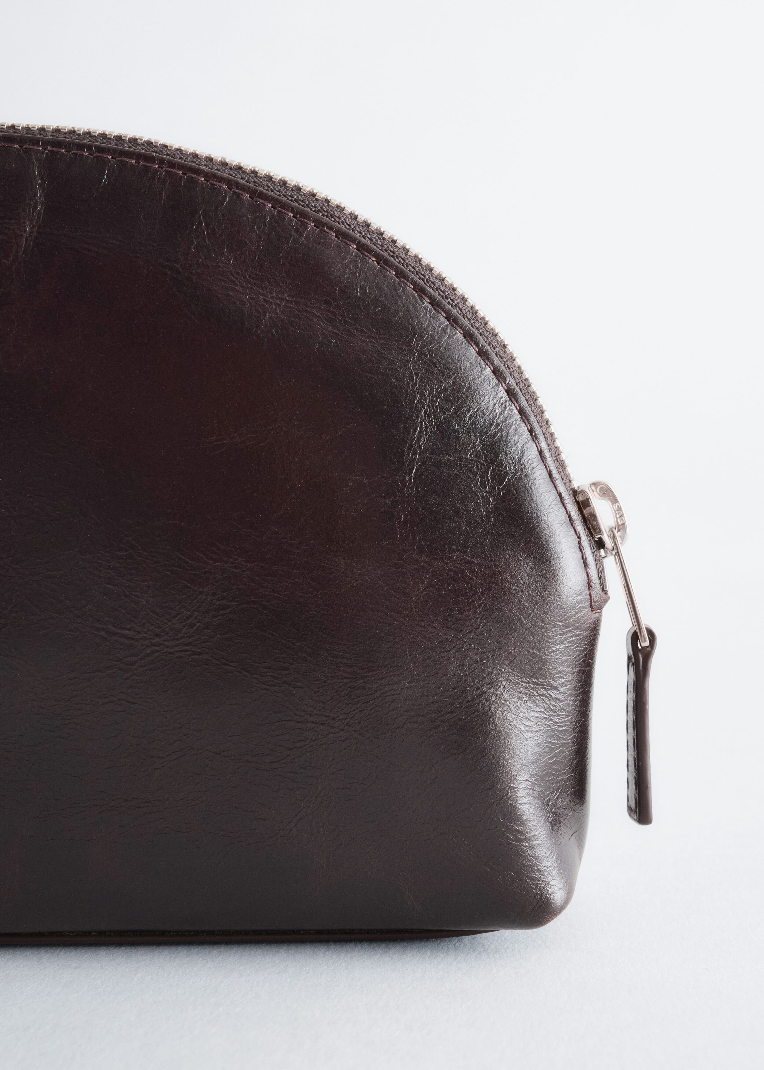 La pochette en cuir brun foncé présente une forme incurvée, en dôme, avec une finition subtilement texturée. Une fermeture éclair en métal ton argent court le long du bord supérieur arrondi, se terminant par une tirette assortie en cuir pendant sur le côté.
