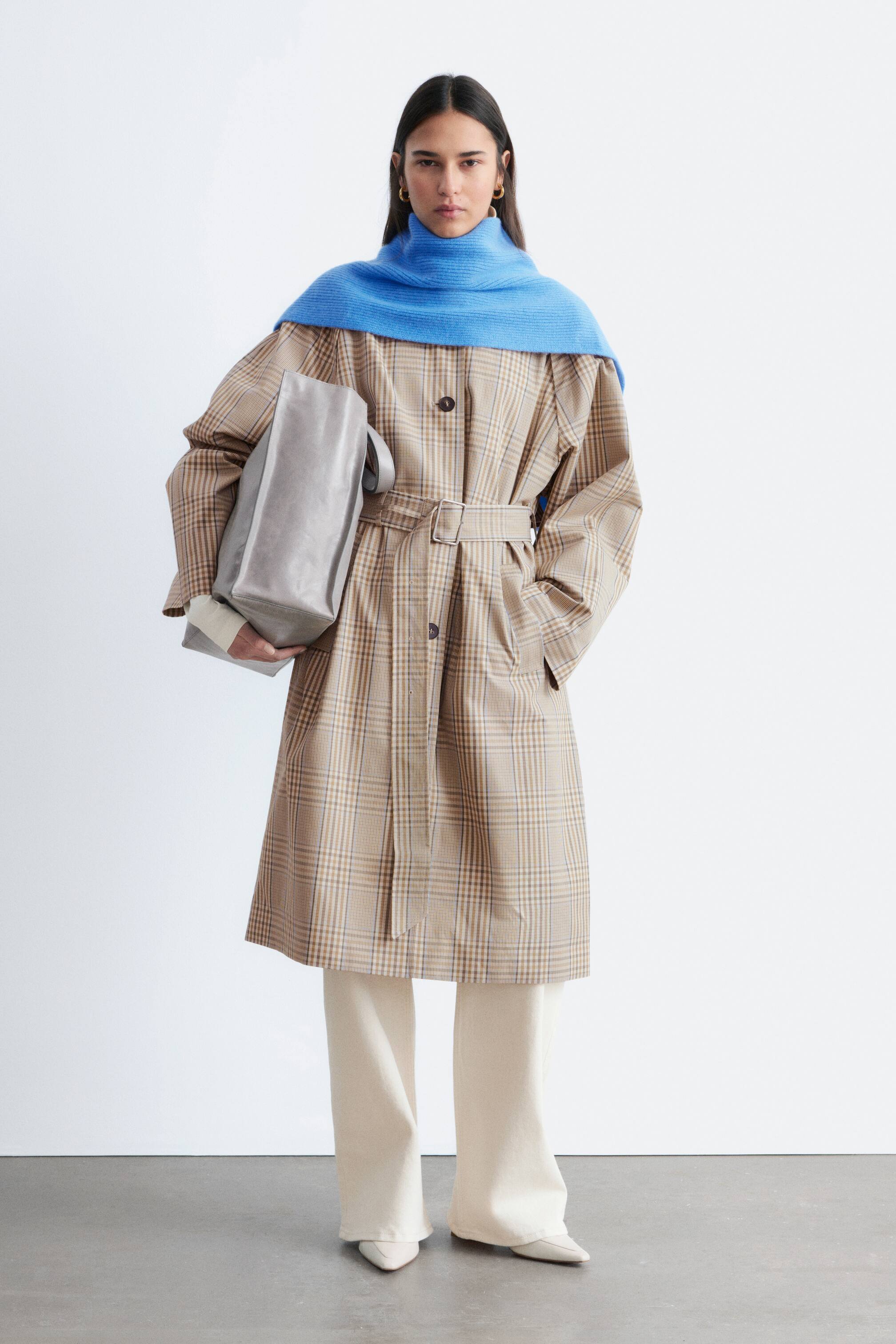 Преглед на по-голямо изображение: Тренчкот с дължина до коленете, обърнат отпред, в бежово-кафяв каре, който се отличава с отпуснат силует, закопчаване с копчета отпред, колан в същия десен и странични джобове. Ярко синьо плетиво с едра плетка е стилизирано върху яката и раменете на палтото.