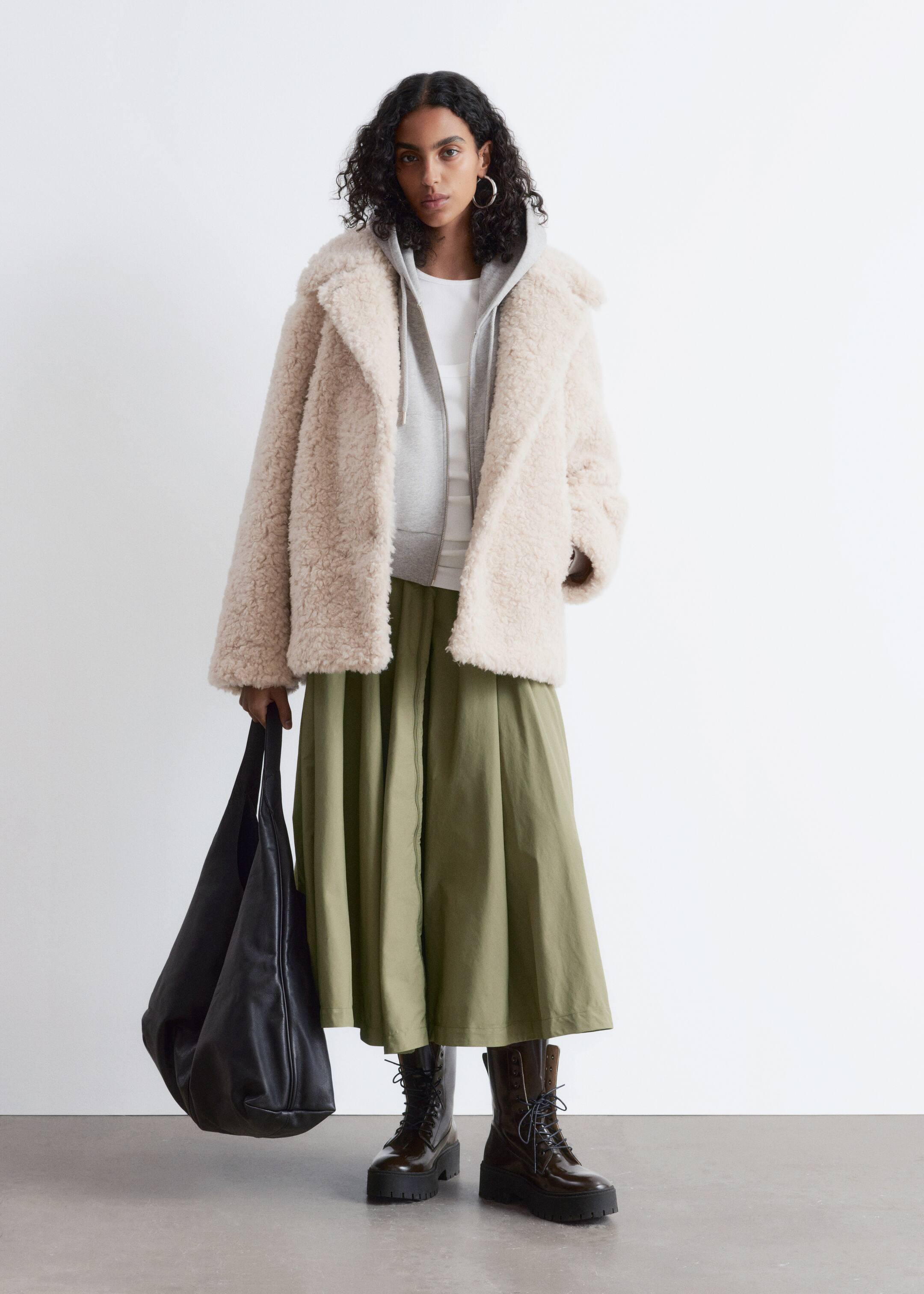 Größeres Bild ansehen: Der beigefarbene Mantel im Shearling-Stil wird an einer Person präsentiert, die leicht nach rechts schaut und die offene Vorderseite zeigt. Er verfügt über breite Kerbrevers, eine strukturierte Oberfläche und eine lockere Passform, mit einem hellgrauen Kapuzenpullover, der darunter sichtbar ist.