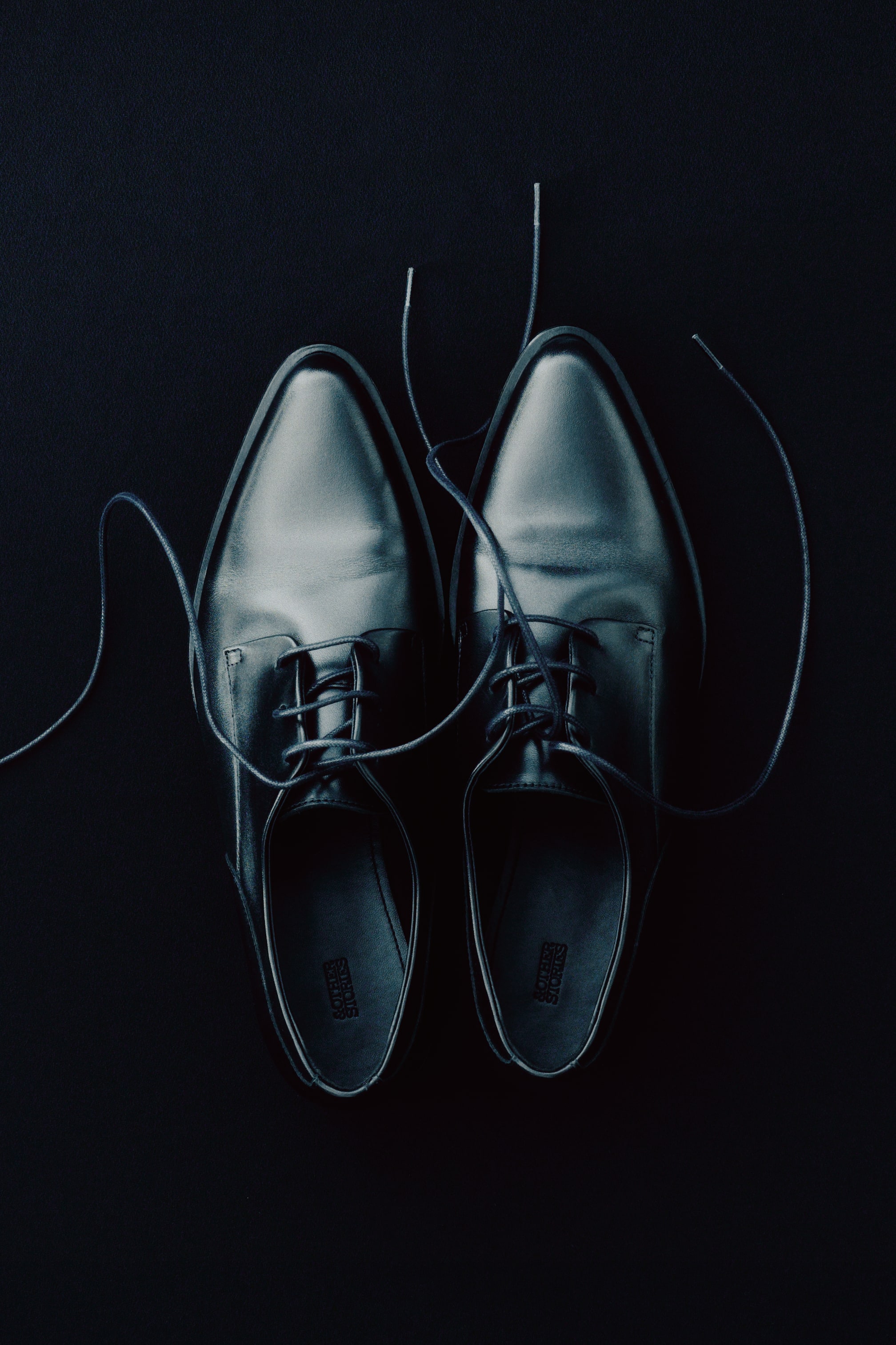 View larger image: Two black leather dress shoes are displayed from a top-down perspective, pointing upwards. Unlaced black shoelaces are casually draped around their sleek design.