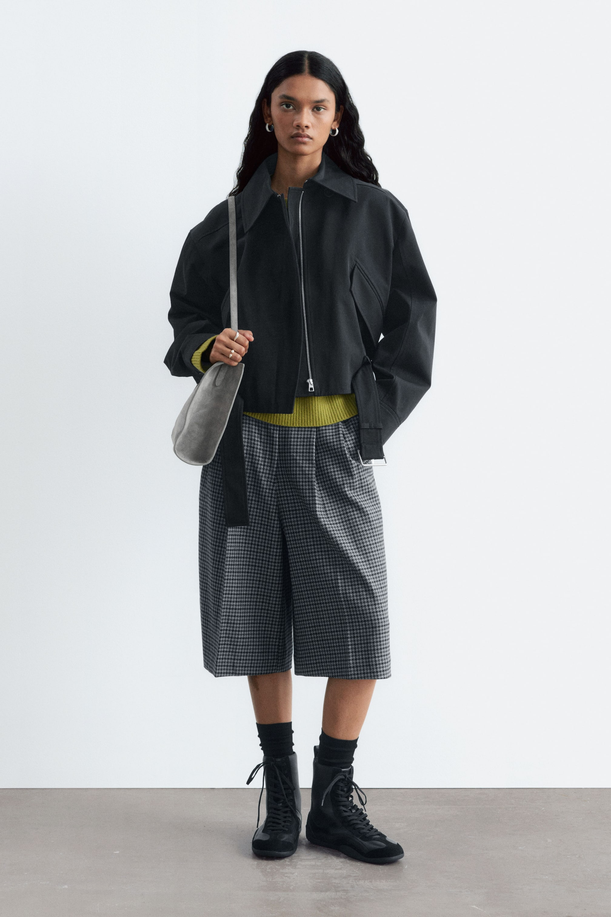 Visualizza immagine più grande: Una giacca bomber nera corta viene indossata aperta sopra un maglione color chartreuse, abbinata a una culotte a quadri grigi. La modella porta una borsa a spalla grigia e indossa stivali neri con allacciatura.