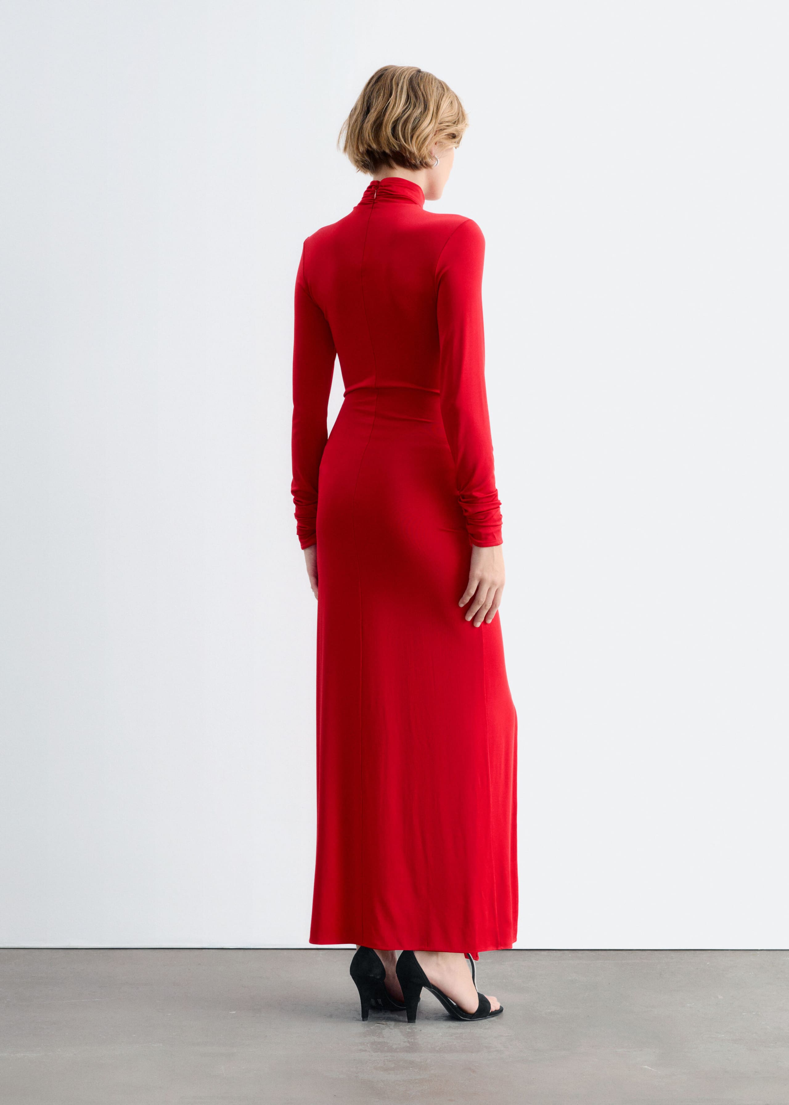 A long-sleeved red jersey maxi dress with a high mock neck is being worn by the model, showing the back view of its slim-fit silhouette. The model is standing with her back to the camera, pairing the ankle-length dress with black high-heeled sandals.