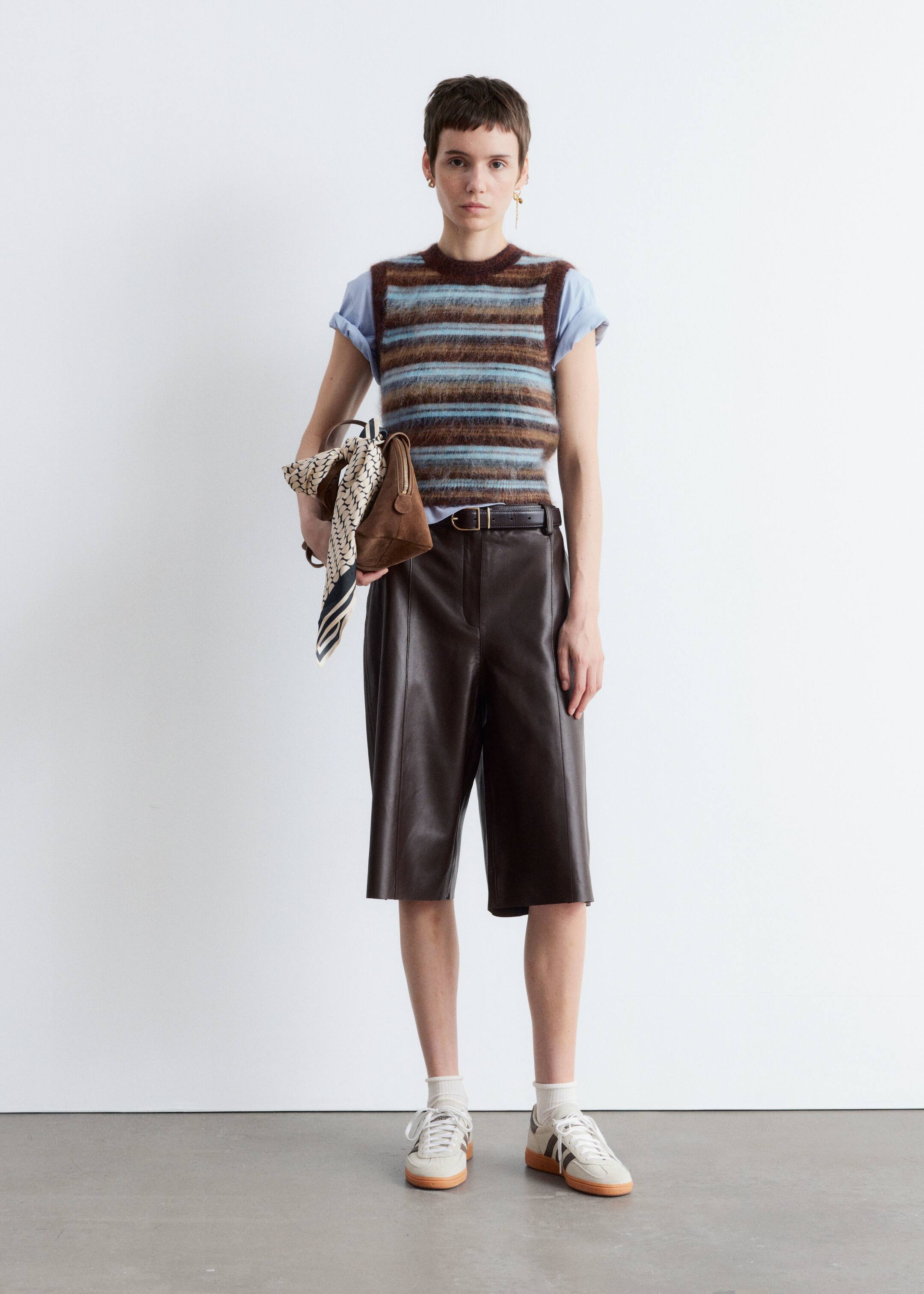 Größeres Bild ansehen: Ein Outfit in voller Länge wird gezeigt, mit einer Person, die nach vorne schaut und eine gestreifte, flauschige Strickweste in Braun- und Blautönen über einem hellblauen T-Shirt mit kurzen Ärmeln trägt. Dunkelbraune Ledershorts mit sichtbaren vertikalen Nähten werden mit einem dünnen braunen Gürtel getragen, während eine kleine braune strukturierte Handtasche, die mit einem gemusterten Schal verziert ist, gehalten wird, und hellfarbene Low-Top-Sneaker mit dunklen Seitenstreifen und weißen Socken vervollständigen den Look.