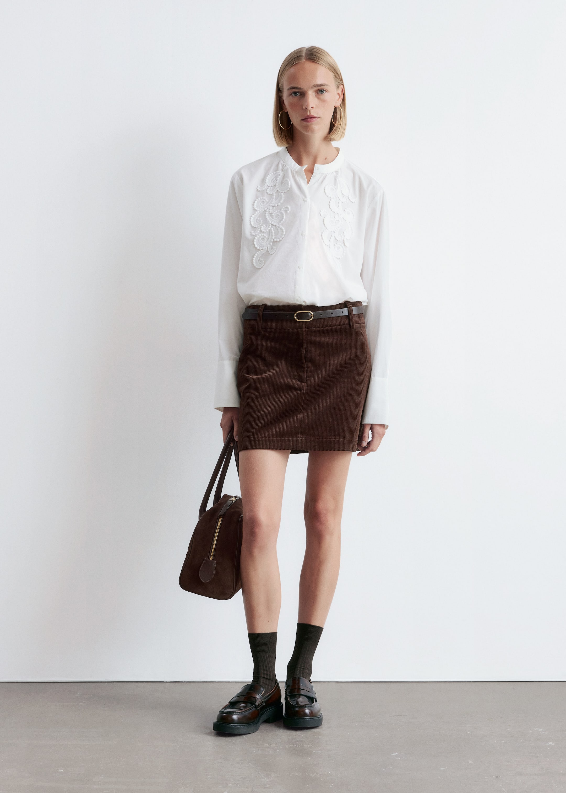View larger image: A white long-sleeved shirt, featuring a band collar and intricate white floral embroidery on the chest, is being worn with a brown corduroy mini skirt, cinched by a thin brown leather belt with a gold-toned buckle. Brown leather loafers with a chunky black sole and a penny loafer strap are being worn over dark ribbed socks, while a brown duffel-style bag is being held.