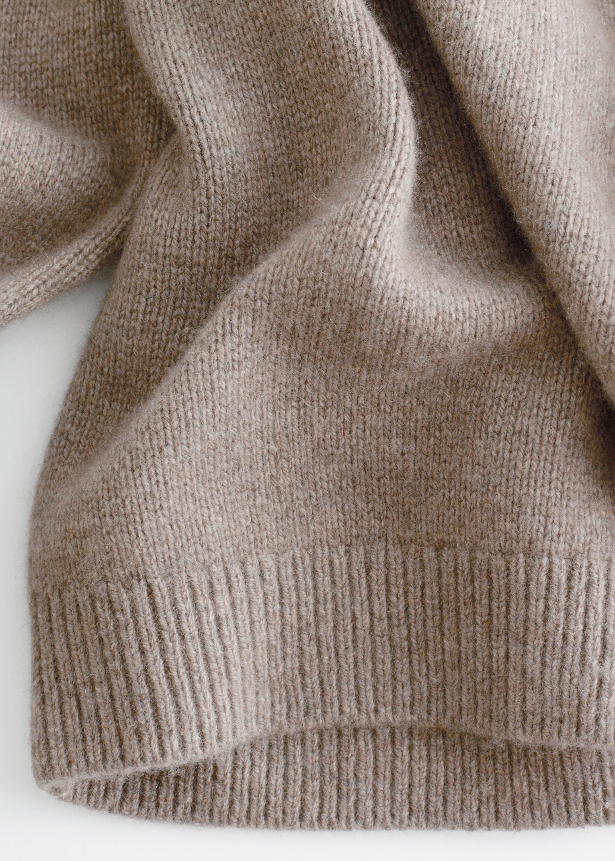 Grösseres Bild ansehen: Ein hellbeiger Strickpullover wird in einer Nahaufnahme präsentiert, die sein weiches, feines Strickmuster und einen markanten gerippten Saum zeigt.