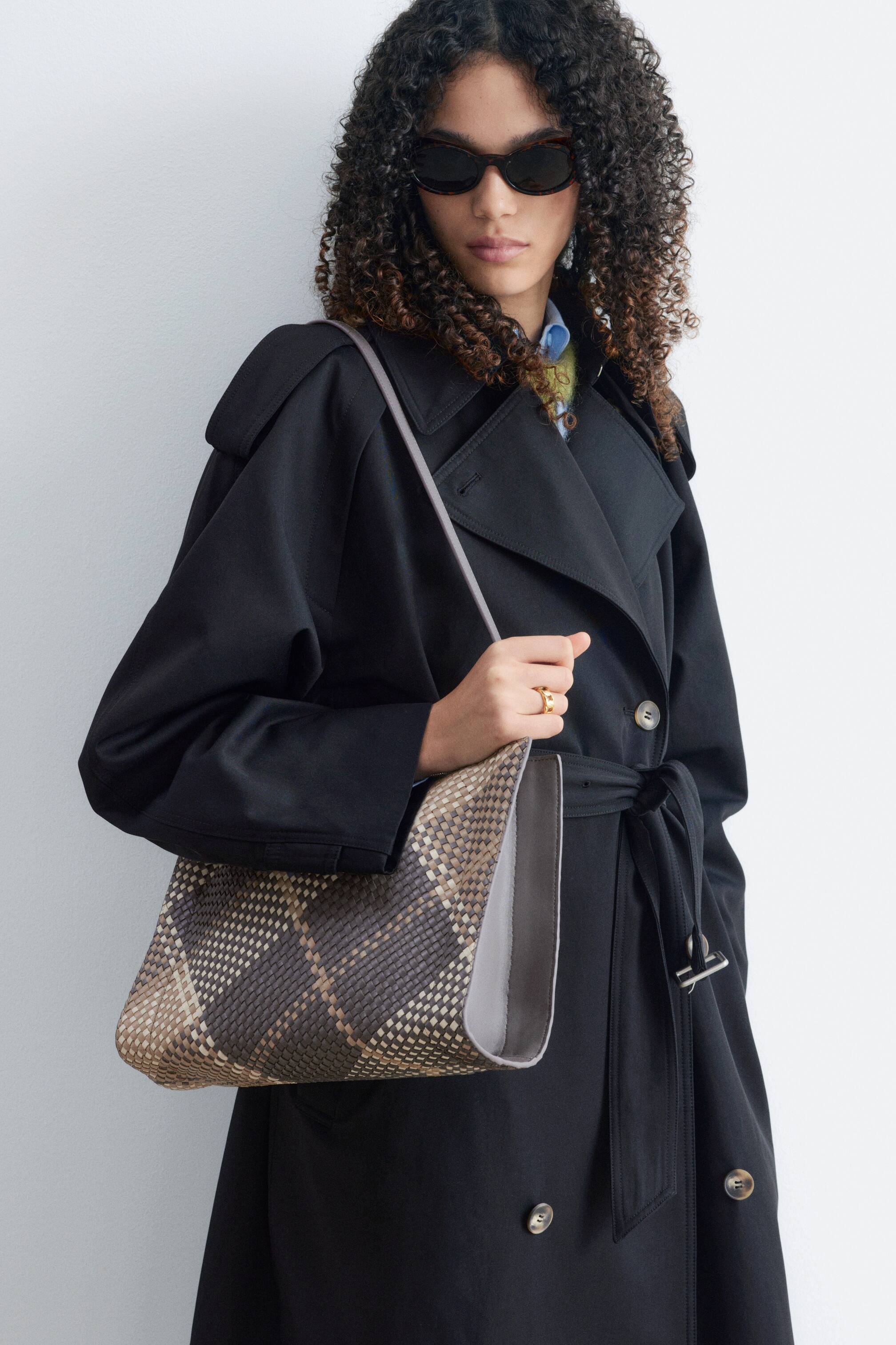 Agrandir l'image: Un sac bandoulière en cuir tressé, présentant un motif losange tissé en marron et beige, est porté par le mannequin par-dessus un trench-coat noir. Le sac pend à son côté, et elle tient sa bandoulière grise de la main droite.