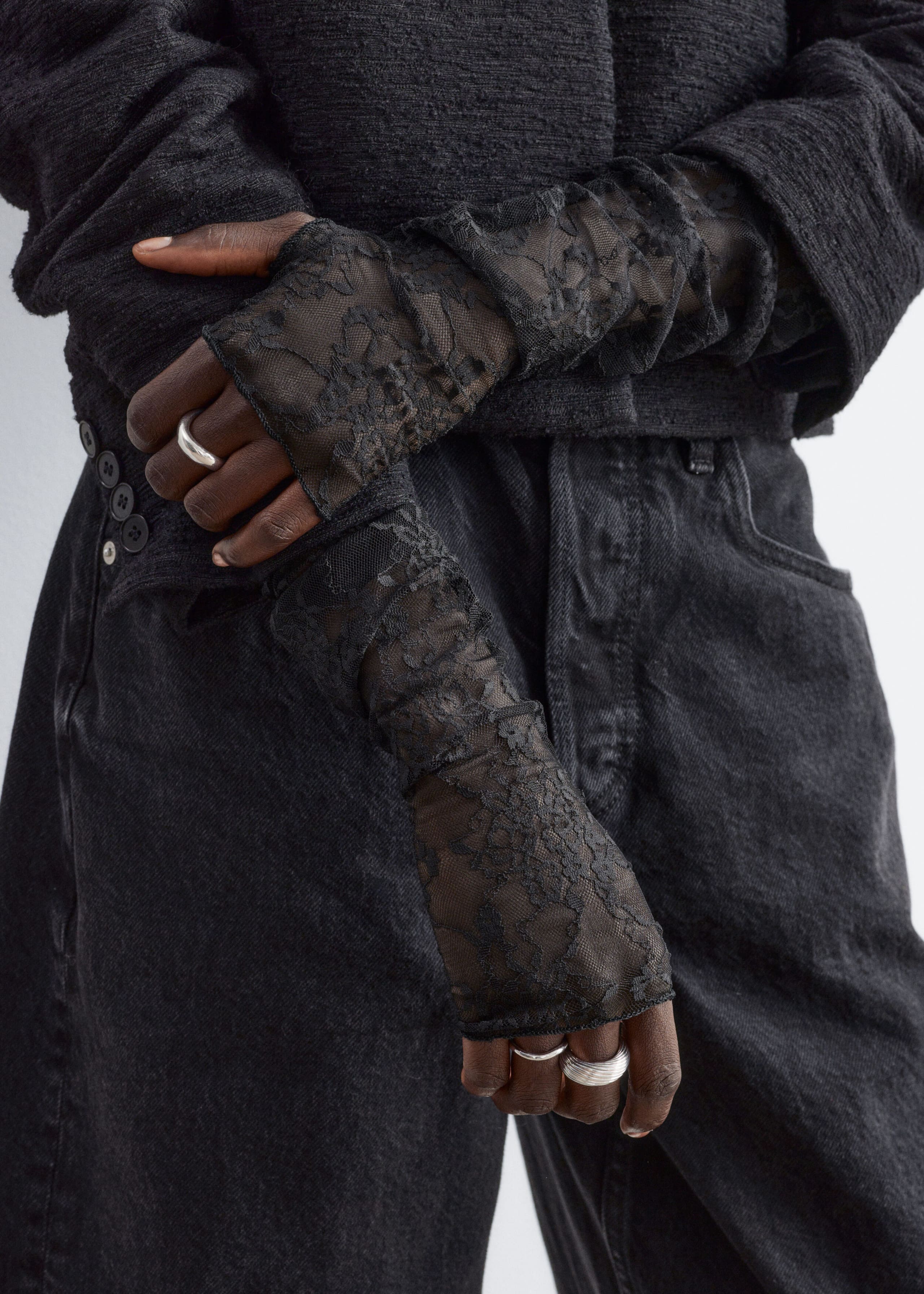 Sheer black lace fingerless gloves featuring a floral pattern are being worn by the model, extending from the knuckles up the forearms. The long, slightly gathered gloves are styled with a textured black jacket, dark denim jeans, and silver rings.