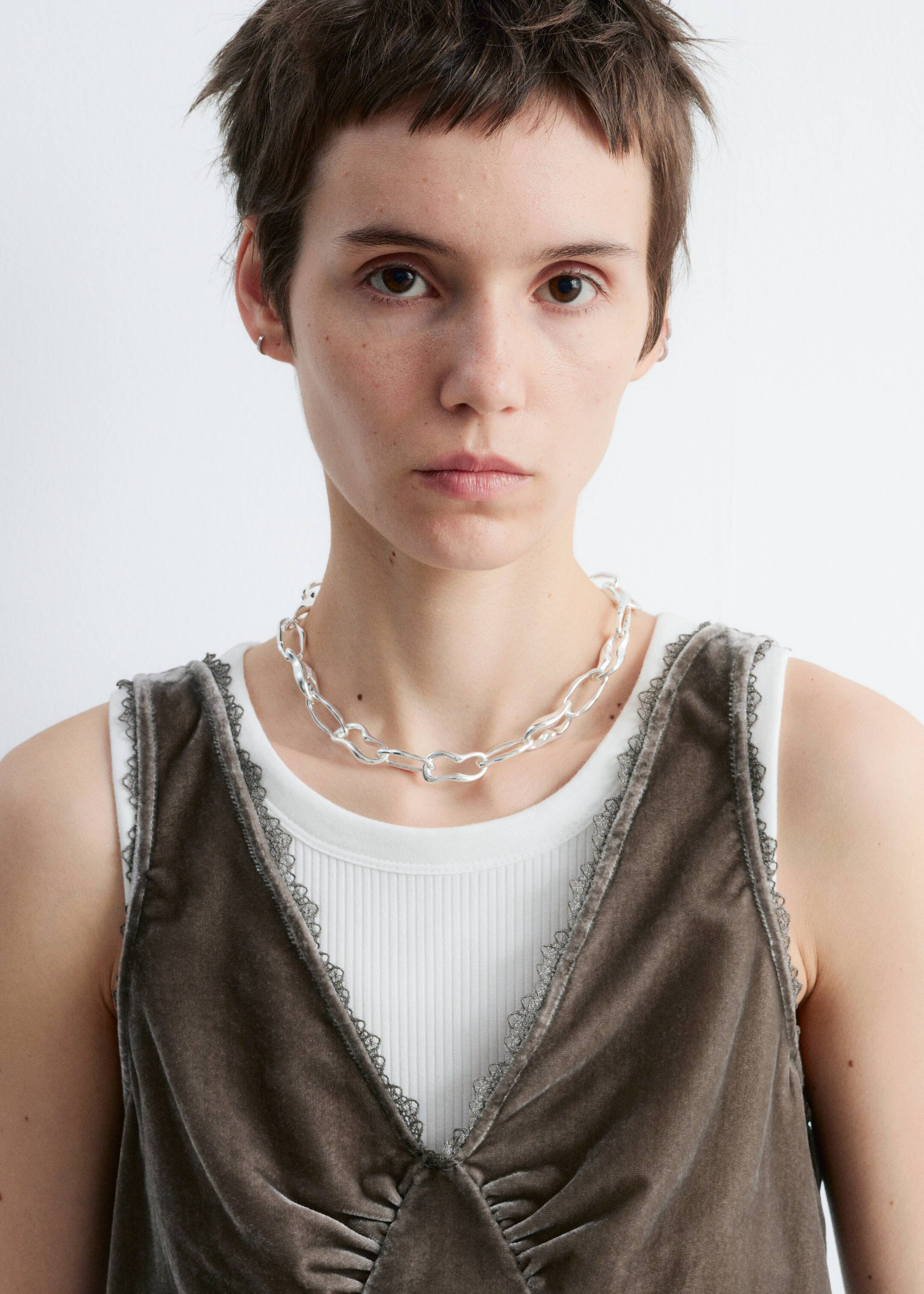 Größeres Bild anzeigen: Eine silberfarbene Kettenhalskette mit großen, unregelmäßigen ovalen Gliedern liegt über einem weißen gerippten Tanktop. Dieses Tanktop ist unter einem dunkelgrauen Samt-Camisole geschichtet, das einen tiefen V-Ausschnitt und Armlöcher mit dunklem Spitzenbesatz aufweist und in der vorderen Mitte gerafft ist.