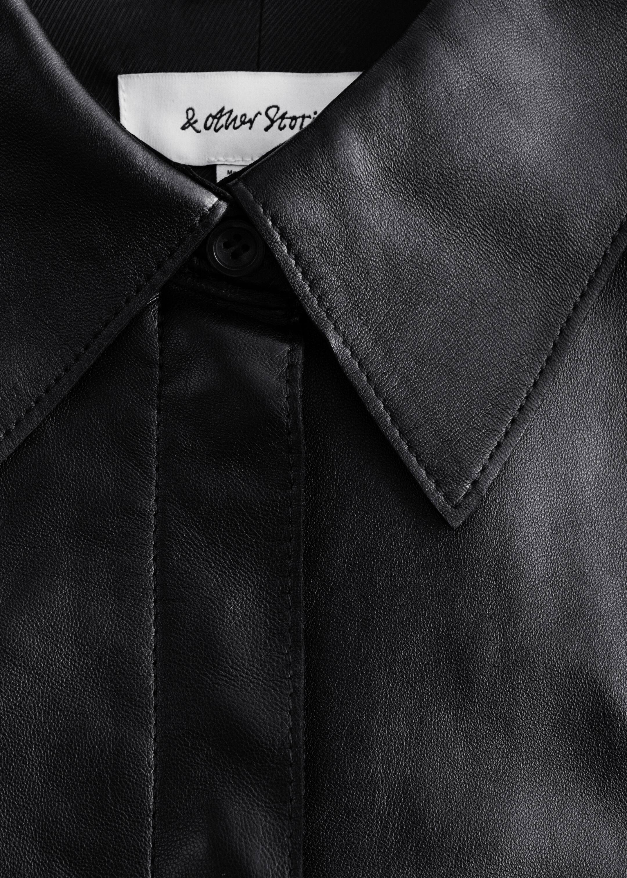 Größeres Bild anzeigen: Ein schwarzes Ledershirt wird in einer Nahaufnahme von vorne präsentiert, mit einem klassischen Kragen mit sichtbaren weißen Nähten und einer verdeckten Knopfleiste mit einem einzelnen oberen Knopf. Ein weißes Markenetikett ist am hinteren Teil des Kragens sichtbar.