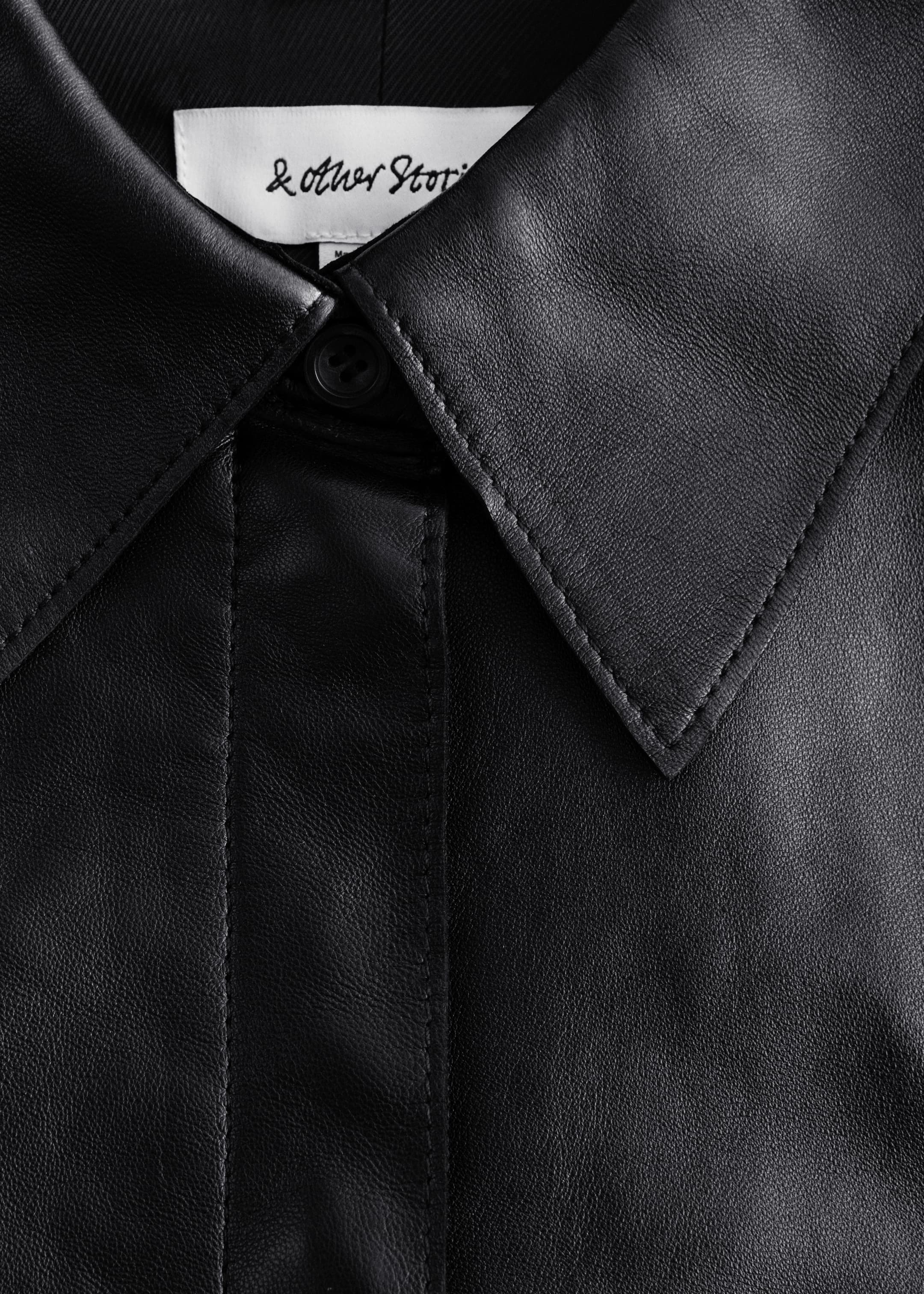 Visualizza immagine più grande: Una camicia in pelle nera viene presentata in una vista frontale ravvicinata, caratterizzata da un colletto classico con cuciture bianche visibili e una lista di bottoni nascosta con un unico bottone superiore. Un'etichetta del marchio bianca è visibile sul retro del collo.