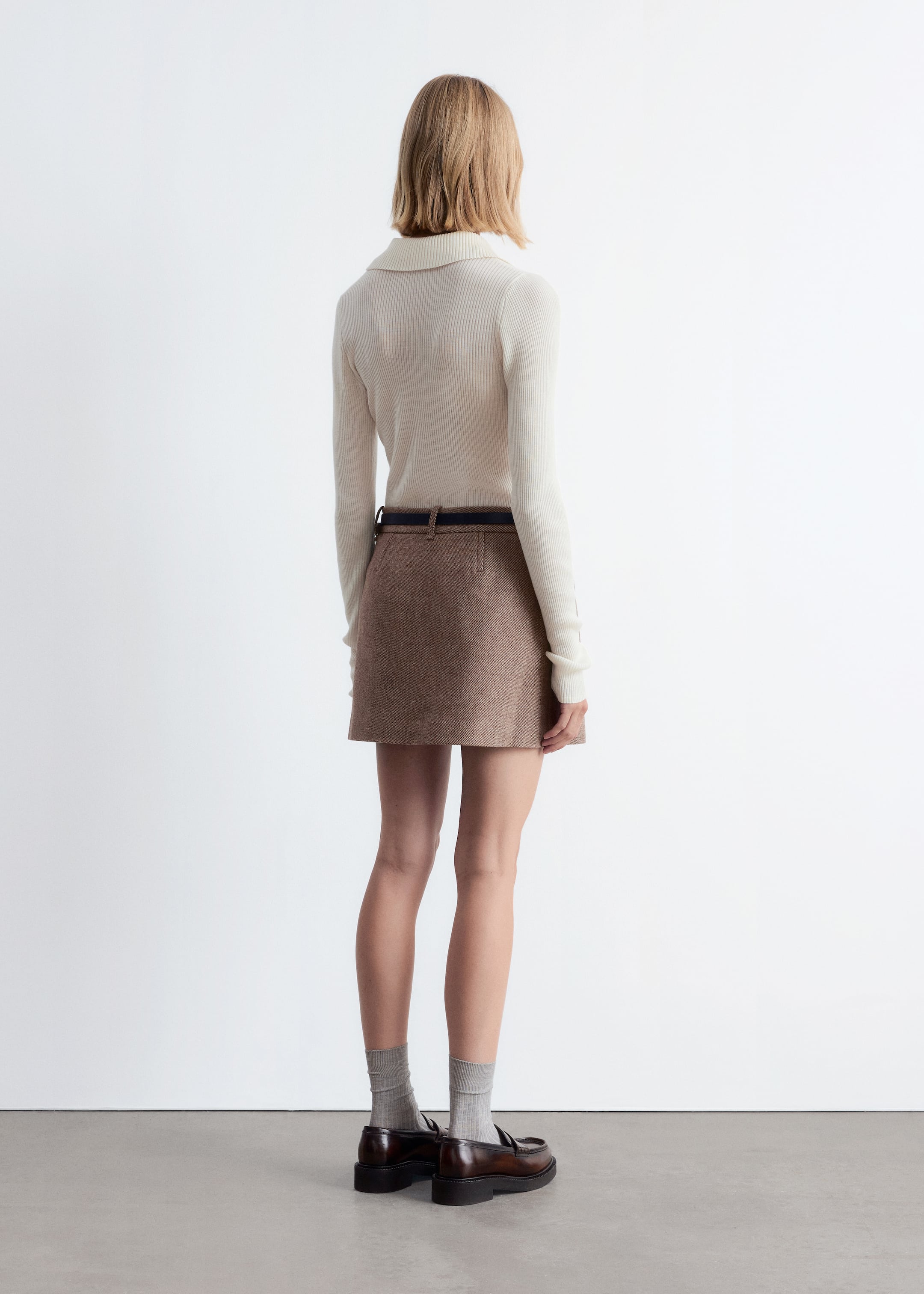 Ingrandisci l'immagine: Un top a maniche lunghe in maglia a coste color crema con un colletto aperto è presentato da dietro. È abbinato a una minigonna in misto lana marrone, caratterizzata da una sottile cintura scura infilata nei passanti.