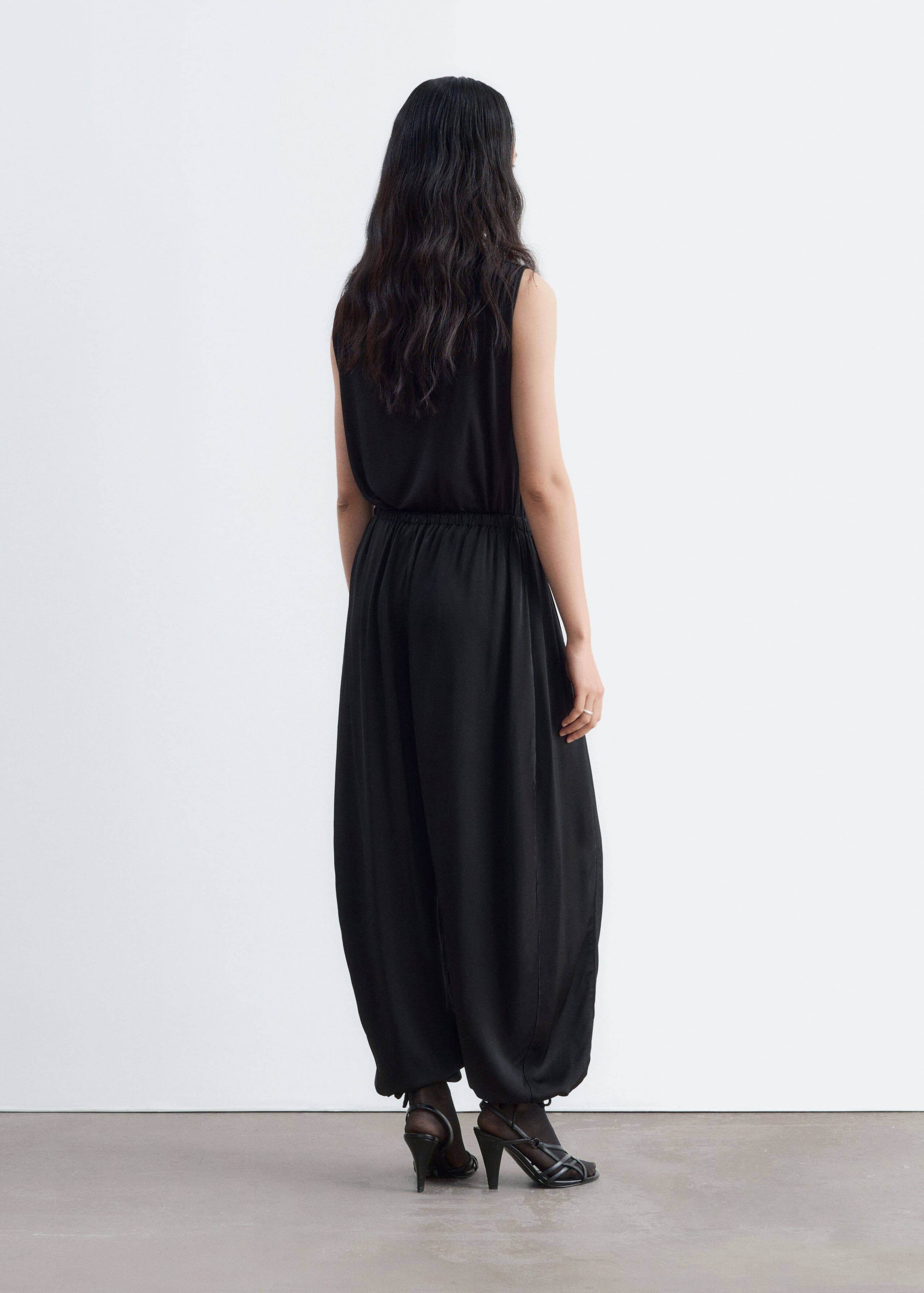 Black balloon-leg trousers made from a fluid fabric are being worn by the model, shown from behind. The loose-fitting trousers feature an elasticated waistband and tied hems, and are styled with a matching black sleeveless top and black strappy high heels.