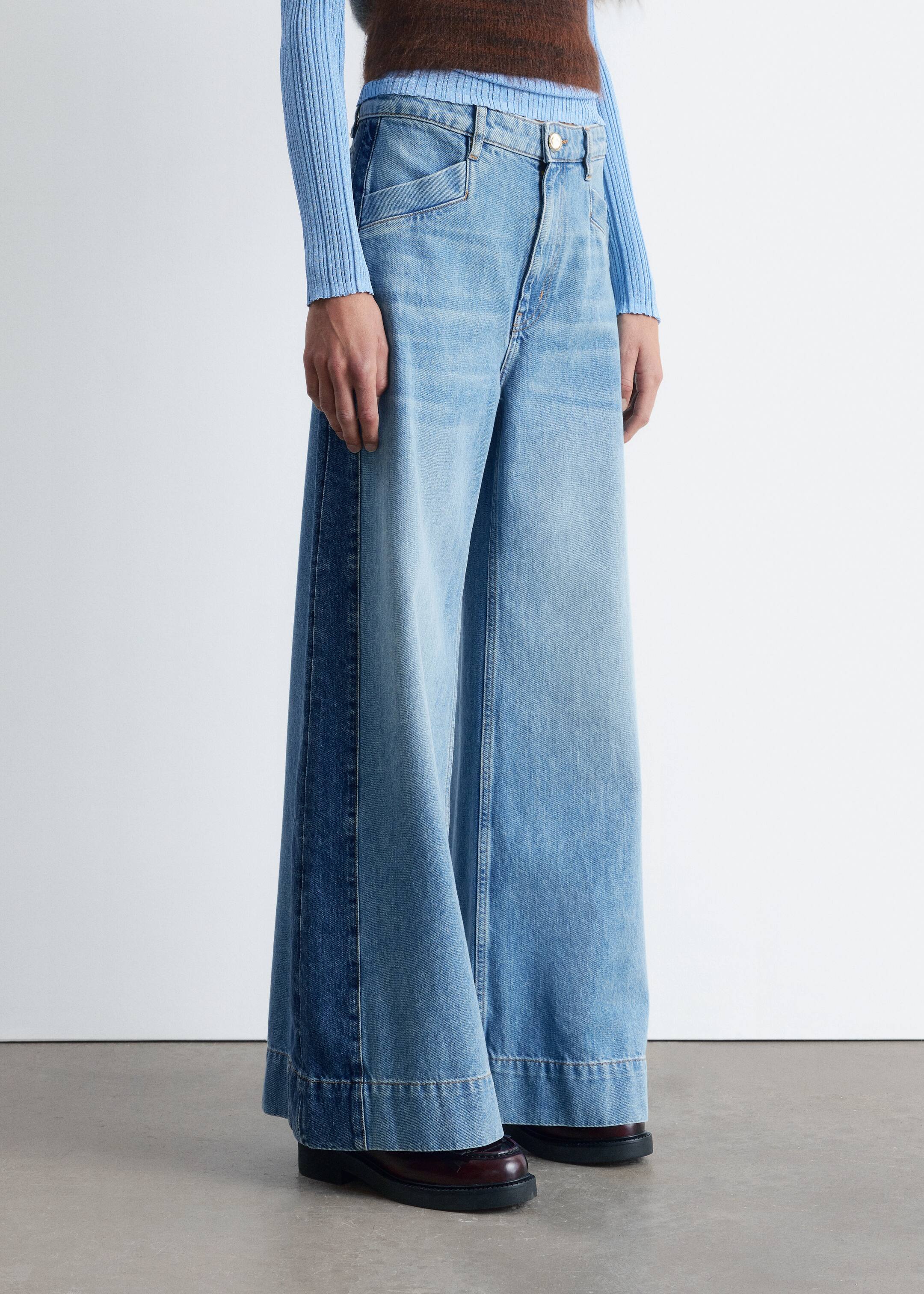 Agrandir l'image: Un jean taille haute, bleu clair avec une silhouette large et longue est porté par le mannequin, qui porte également un haut bleu clair. Le jean présente des détails de panneaux latéraux plus foncés et un ourlet large surpiqué, avec la longueur supplémentaire s'accumulant sur des chaussures sombres.