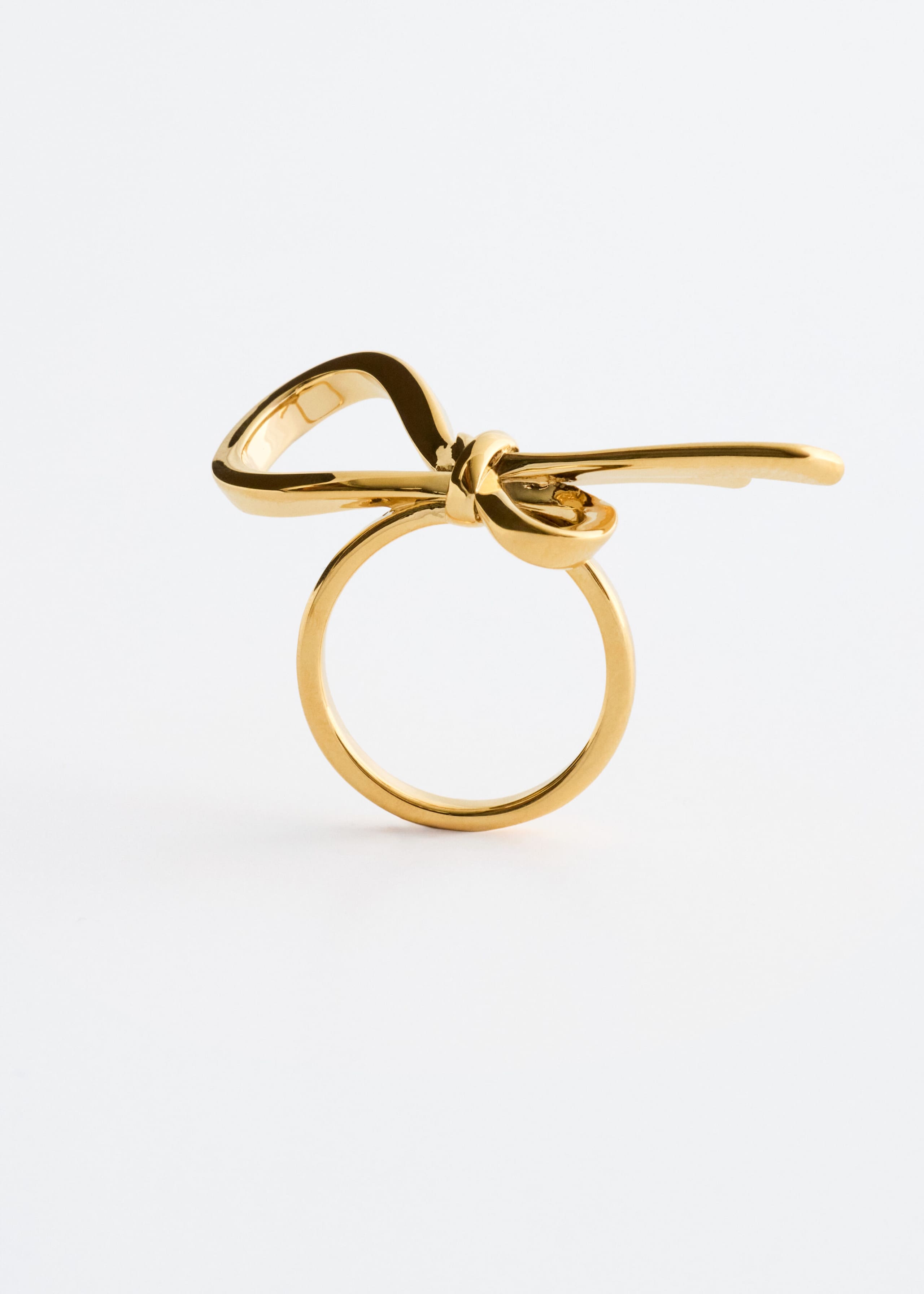 A polished brass ring stands upright, facing forward, showcasing a sculptural knot design shaped like an asymmetrical bow. The ring features a highly reflective metallic finish, with a single loop on the left and a long, smooth tail extending to the right.