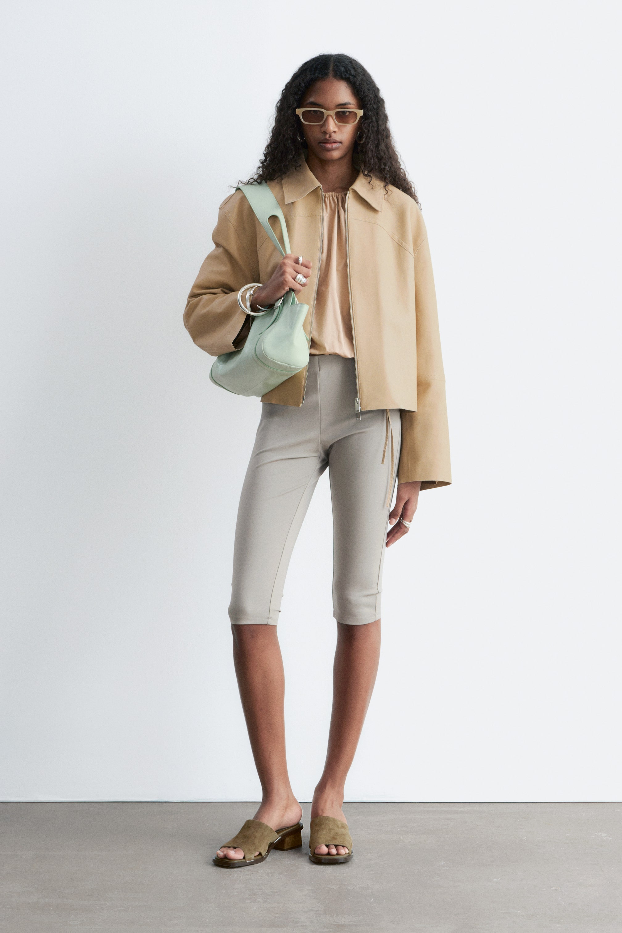 Visualizza immagine più grande: Pantaloni capri beige chiaro, caratterizzati da una silhouette aderente e corta che termina sotto il ginocchio, indossati dalla modella. Presentano una sottile cucitura verticale su ciascun lato esterno e una decorativa cerniera al fianco destro, abbinati a una giacca marrone chiaro e sabot verde oliva.