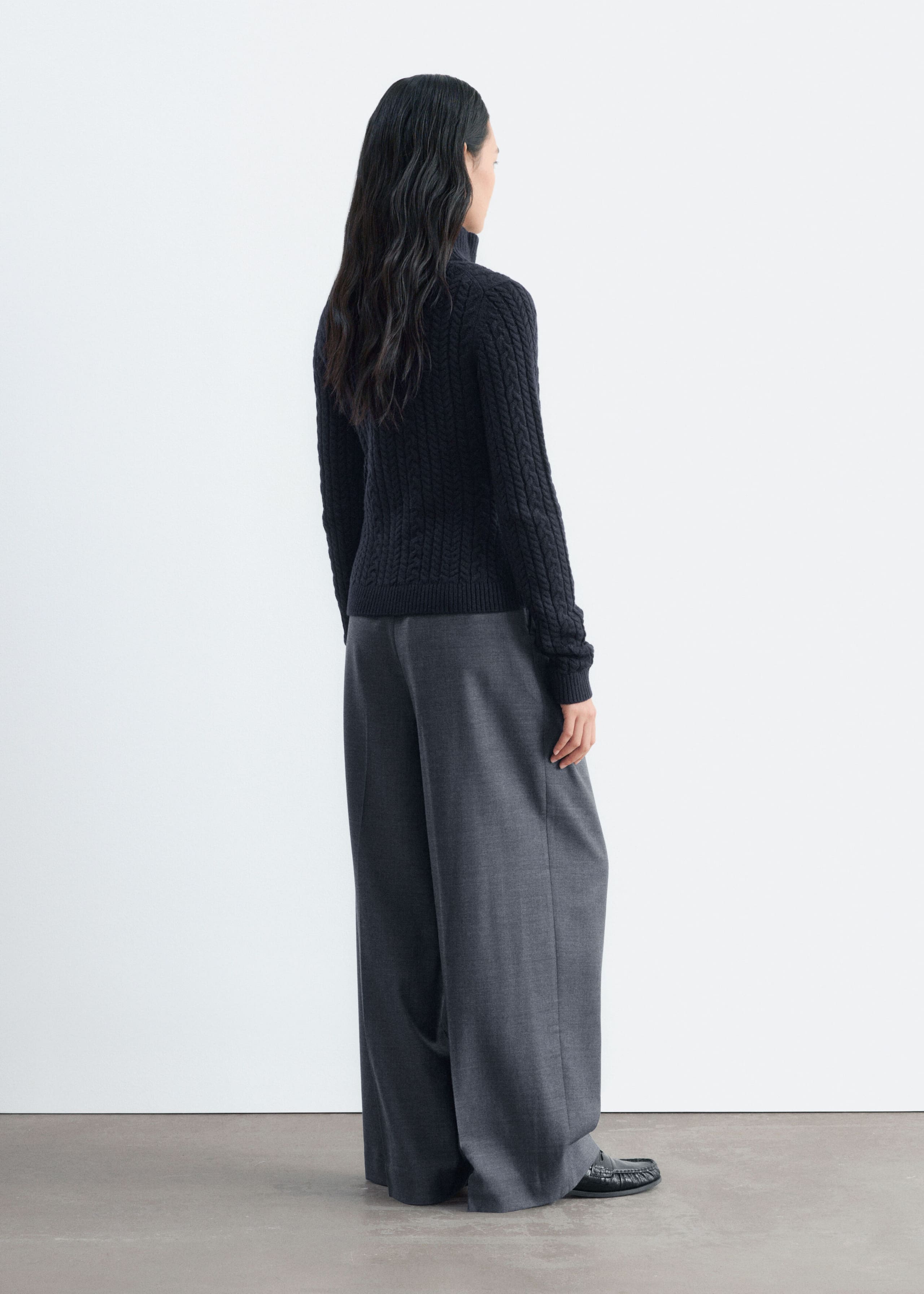 A dark navy cable-knit cardigan is being worn by the model, who is standing in a three-quarter back view. The slim-fit cardigan features a high collar with ribbed cuffs and hem, and is styled with wide-leg grey trousers.