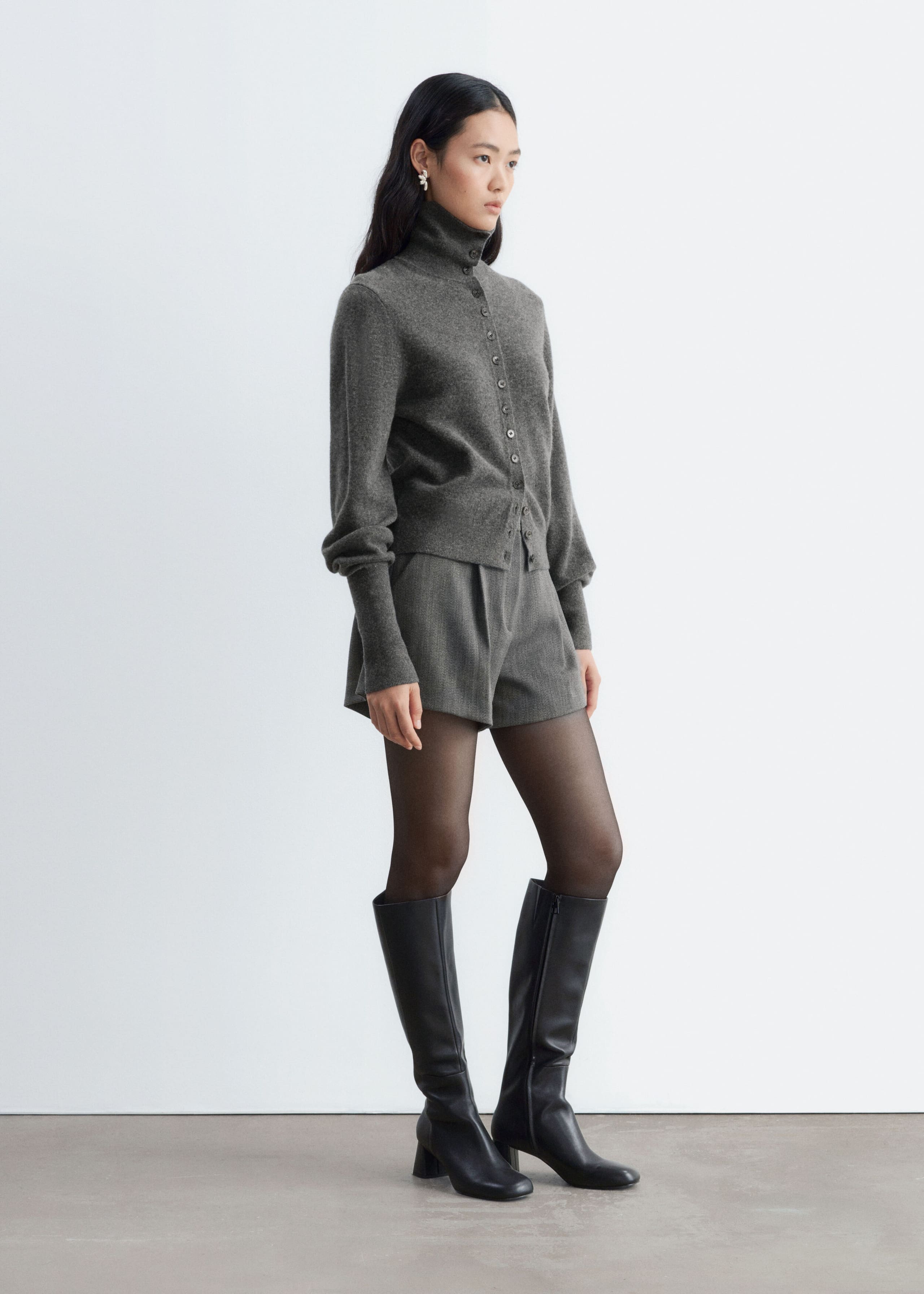 These high-waisted wool-blend shorts in grey, with front pleats and a relaxed A-line silhouette, are worn by the model. They are styled with a matching grey button-up high-neck cardigan, sheer black tights, and knee-high black leather boots.