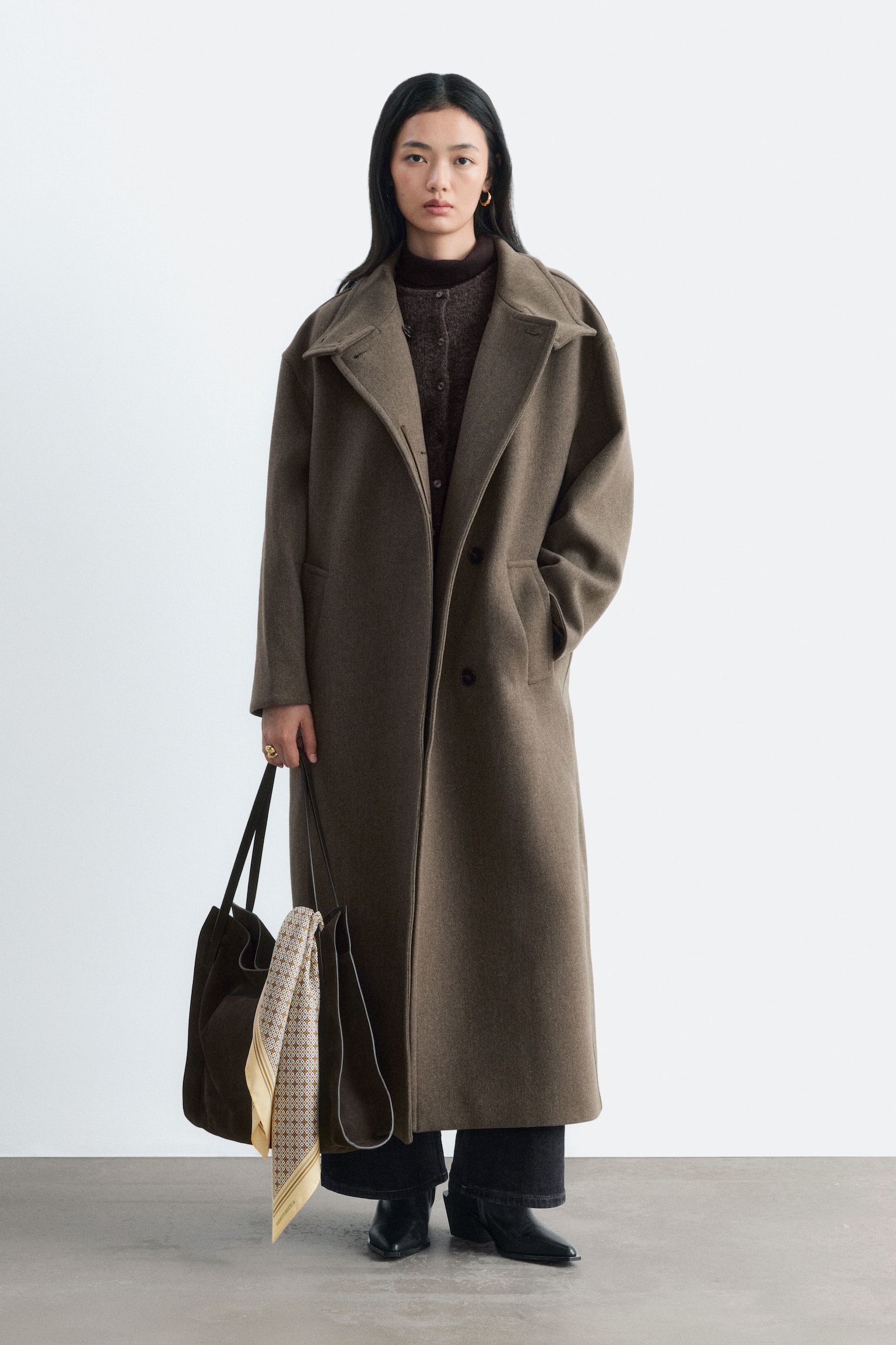 Um casaco de lã oversized, até aos tornozelos, numa tonalidade castanho-oliva escura, está a ser usado pela modelo, apresentando lapelas largas com entalhe e bolsos laterais. O casaco é combinado com um cardigan castanho-escuro de abotoar e um top de gola alta, com a modelo a manter uma mão no bolso do casaco enquanto transporta uma tote bag escura. - 1