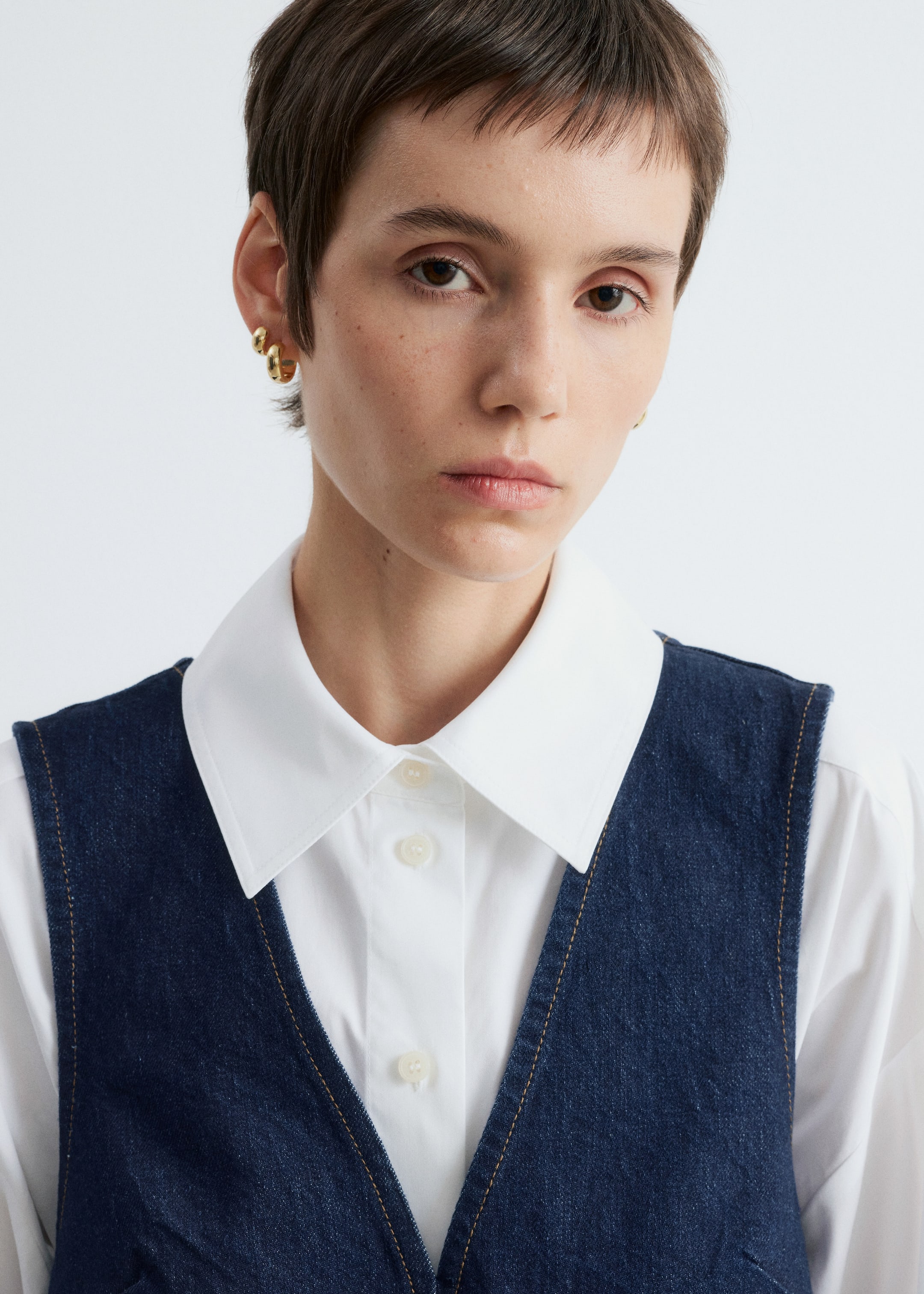 Ingrandisci l'immagine: Un gilet in denim scuro con scollo a V è sovrapposto a una camicia bianca con colletto. I bordi del gilet sono accentuati con cuciture marrone chiaro a contrasto, e il colletto a punta della camicia e i bottoni frontali sono visibili sotto.