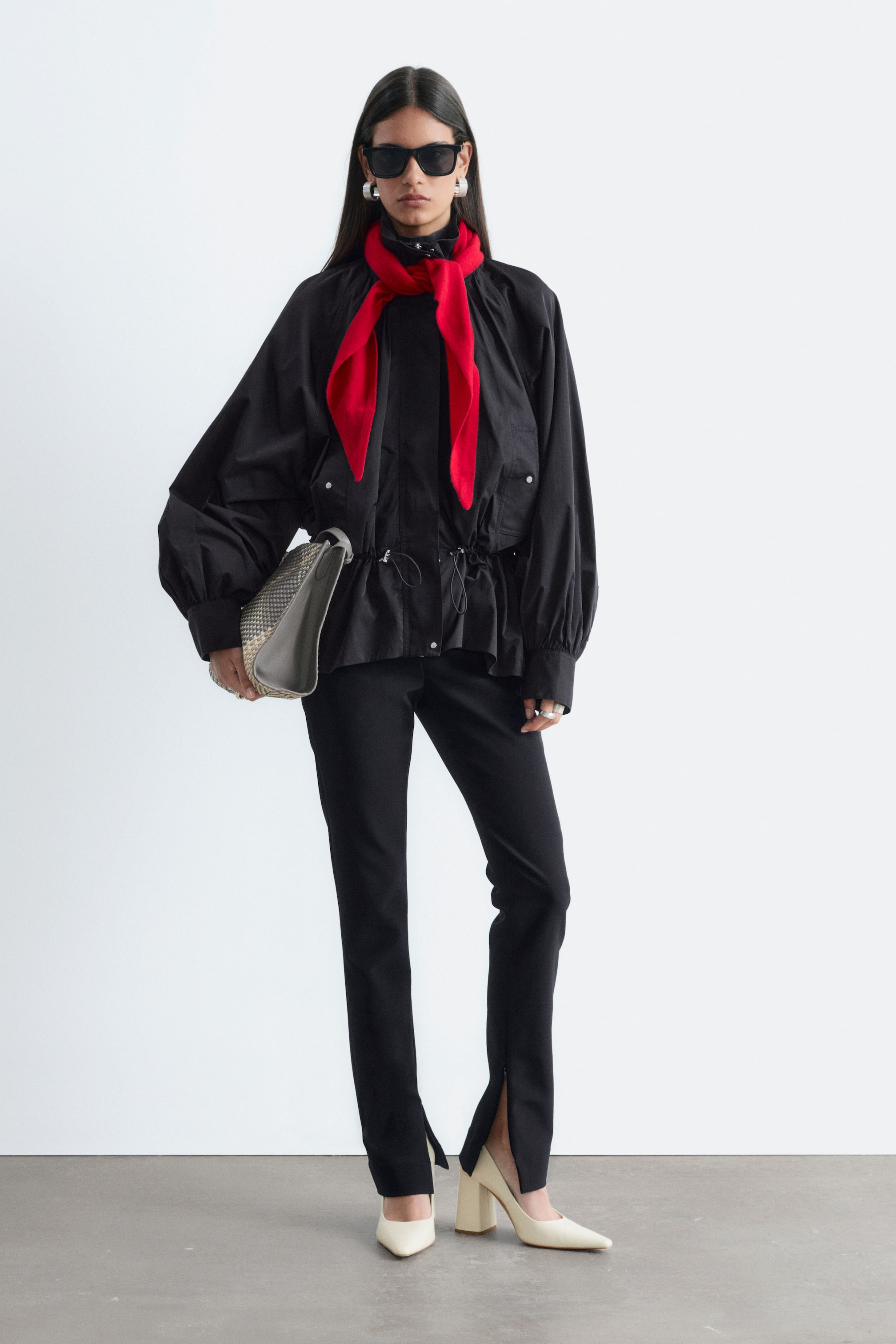 Agrandir l'image: Le mannequin porte une veste en nylon noire à coupe ample, dotée d’un col montant, de manches volumineuses et d’un cordon élastique réglable à la taille froncée. La veste est associée à une écharpe rouge vif enroulée autour du cou, un pantalon noir et une pochette grise portée sous le bras.